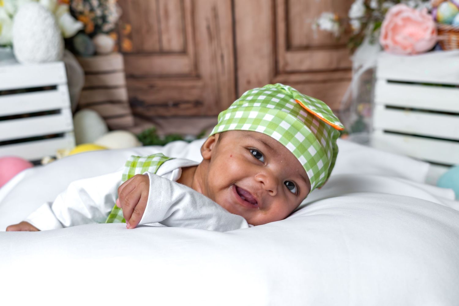 Smiling newborn baby wearing a green checkered hat lying on a white blanket in a festive Easter-themed setting.
