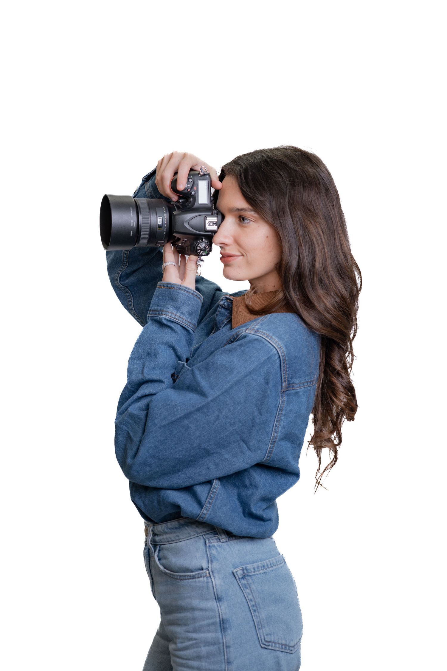 Side profile of young woman in denim outfit holding professional camera up to photograph subject.