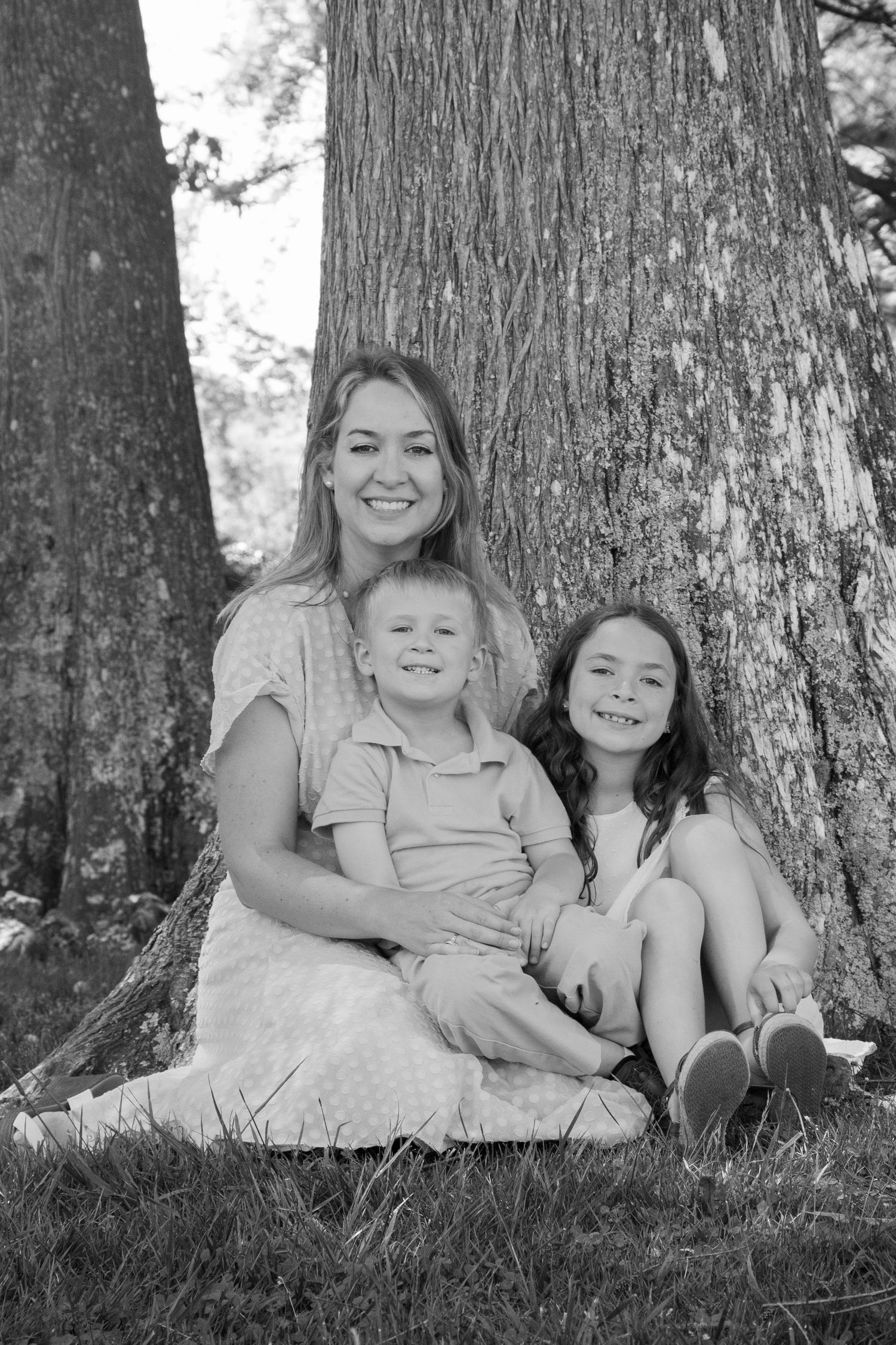 Black and white portrait of smiling mother sitting with two children against large tree trunk outdoors.