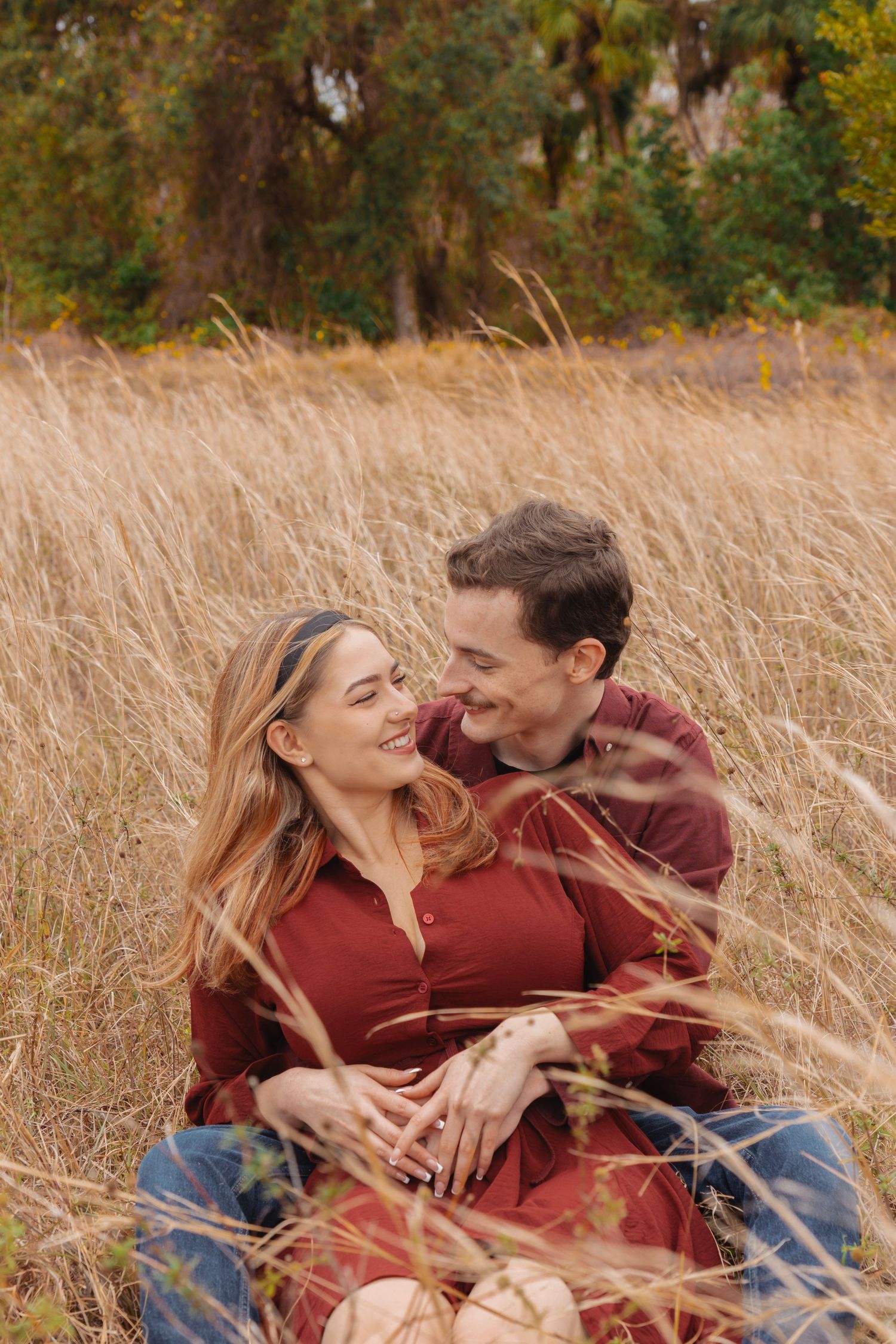 A couple sits in a golden grass field, smiling at each other, wearing matching burgundy outfits in an autumn setting.