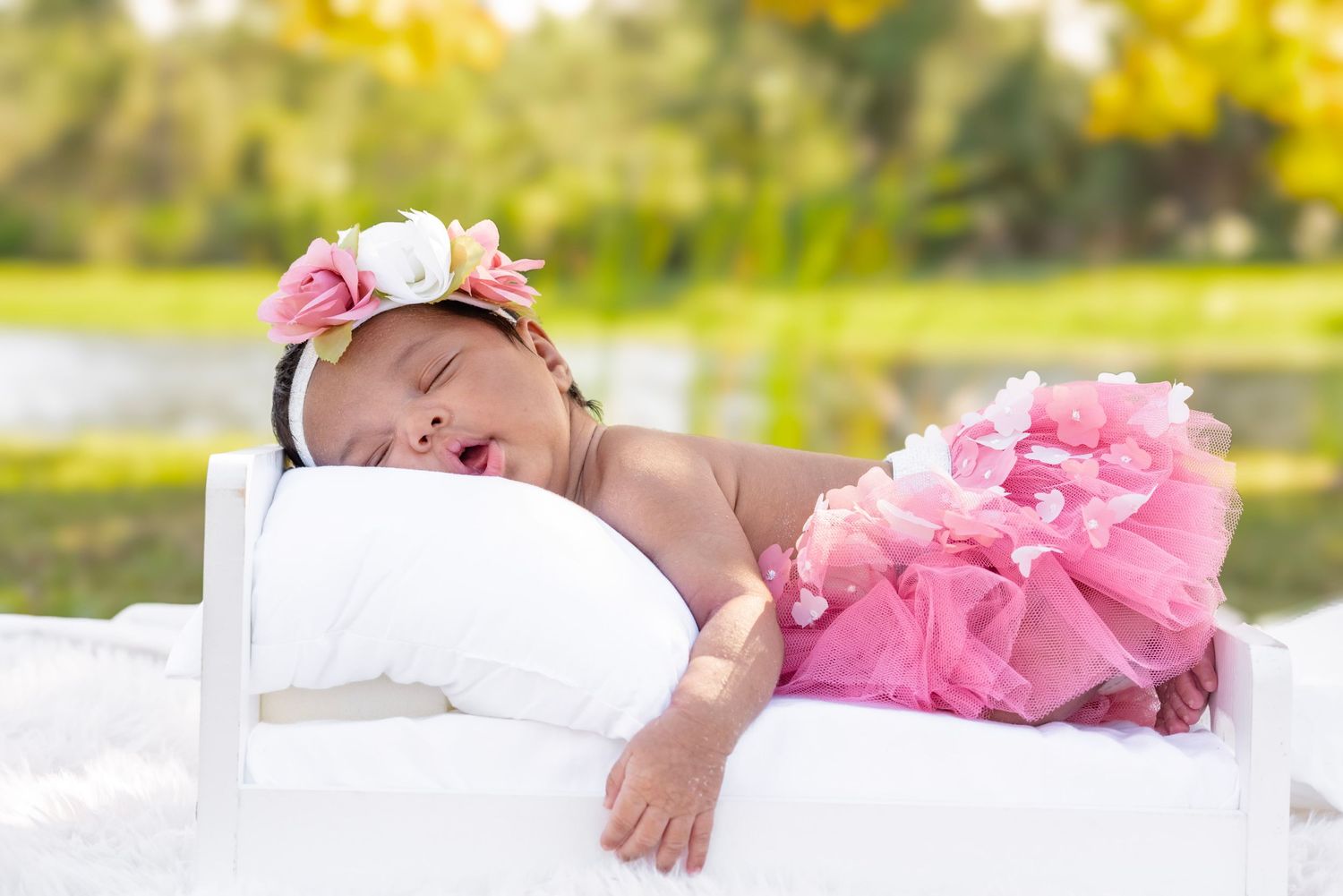 Newborn baby girl yawning in pink tutu skirt and floral headband, sleeping on white pillow outdoors.