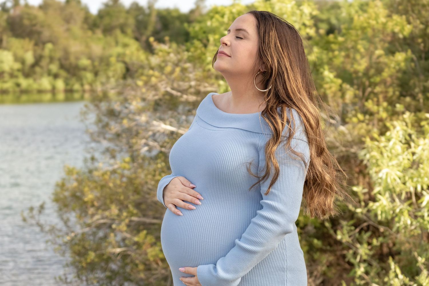 Pregnant woman in blue sweater cradling baby bump outdoors near lake surrounded by lush green trees on sunny day.