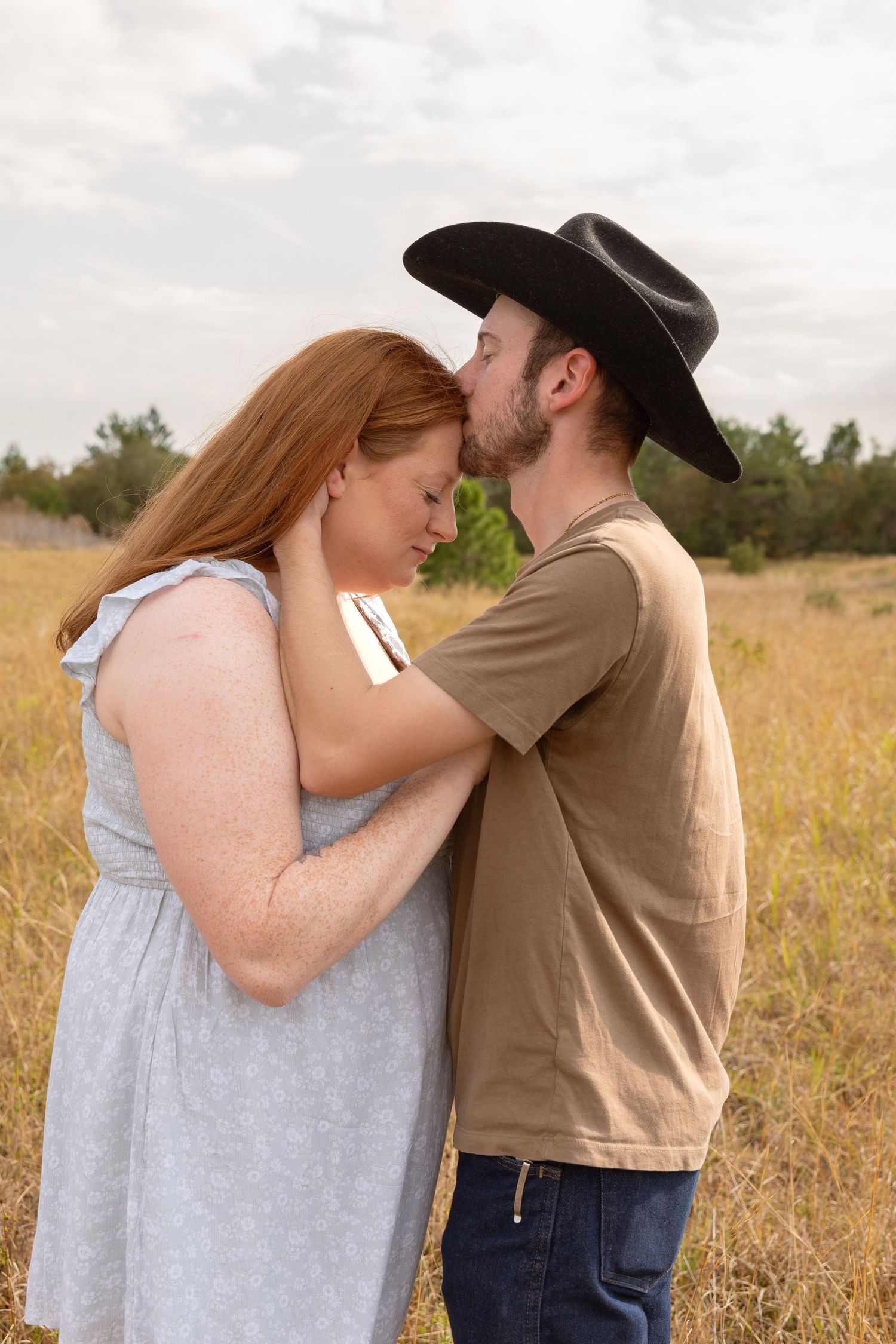 A man in a black cowboy hat kisses a red-haired woman in a blue dress in a golden field.