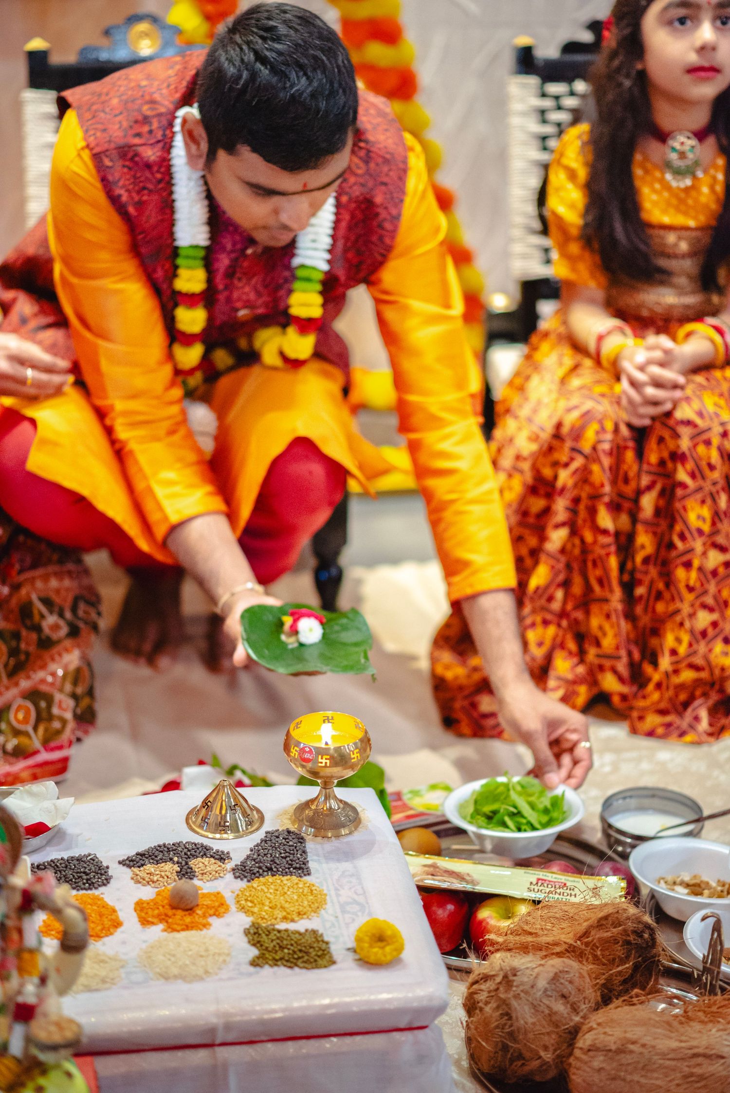 Groom in yellow kurta performs Hindu wedding ritual with grains, flowers, and sacred items on white cloth.