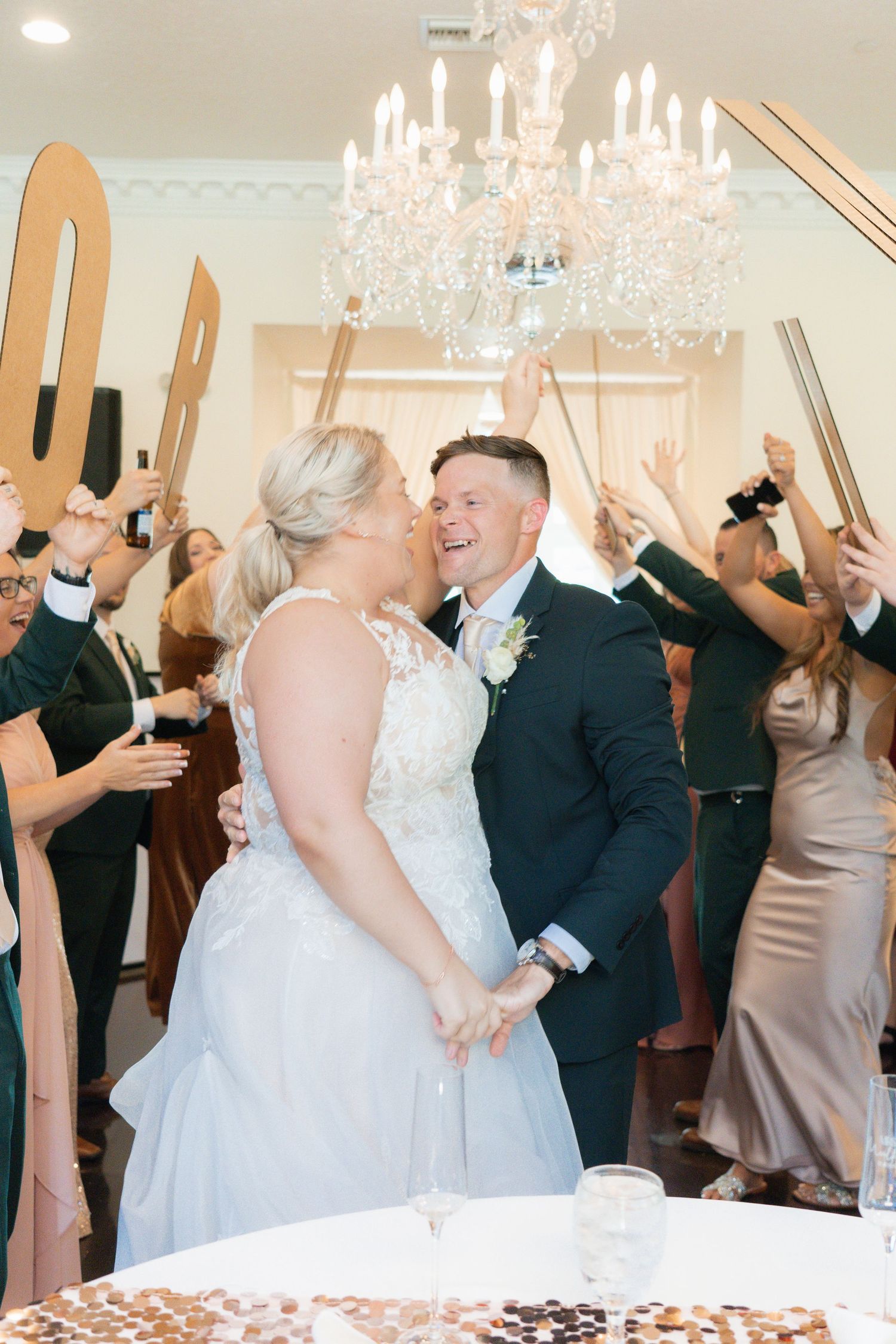 Bride and groom share a kiss during grand reception entrance under crystal chandelier with cheering guests.