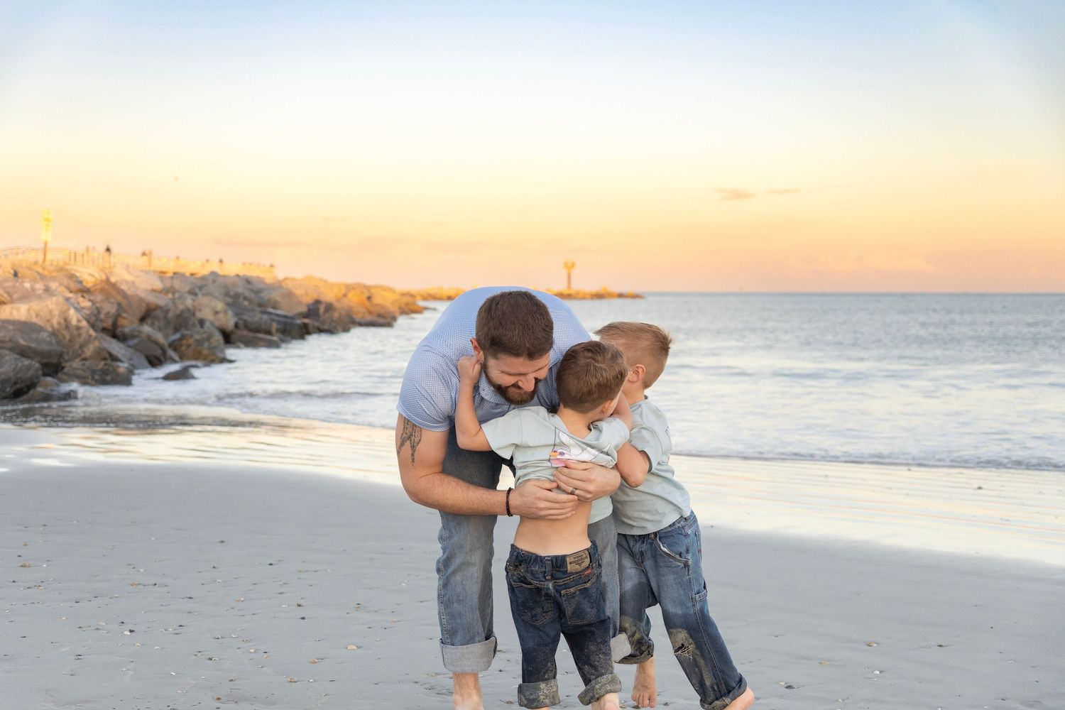 Father hugging two young sons on a sandy beach at sunset with ocean waves and rocky jetty in background.