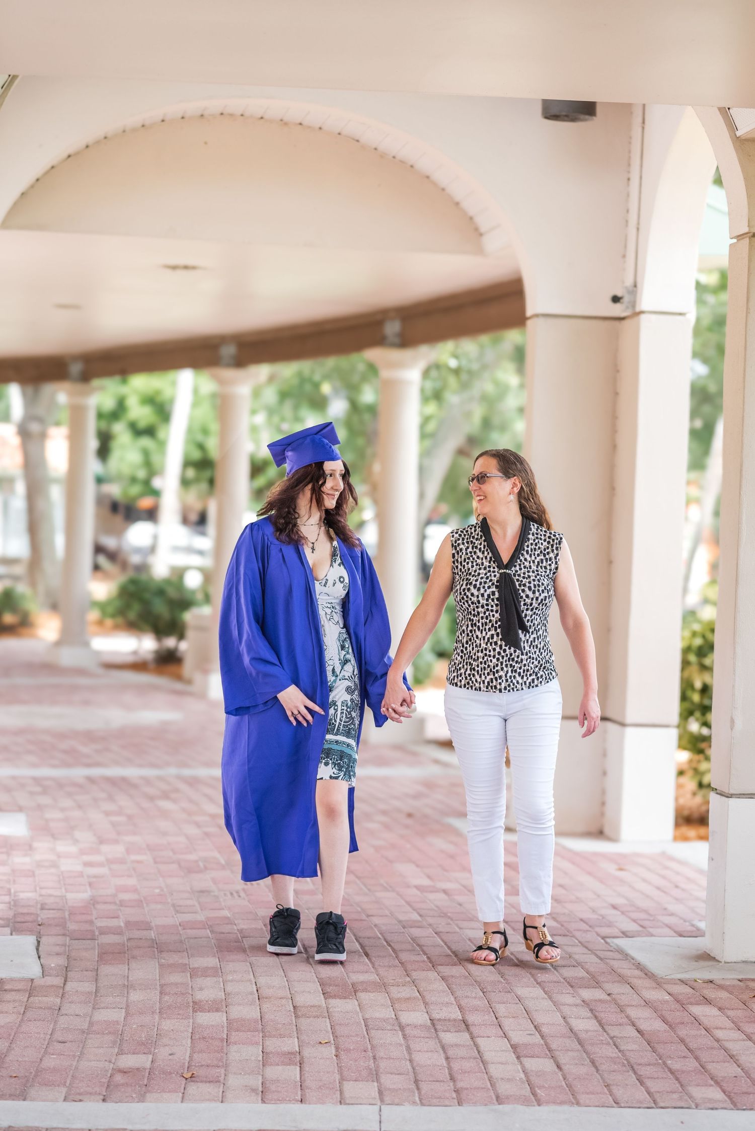 Graduate in blue cap and gown walks hand-in-hand with woman in white jeans under arched colonnade.