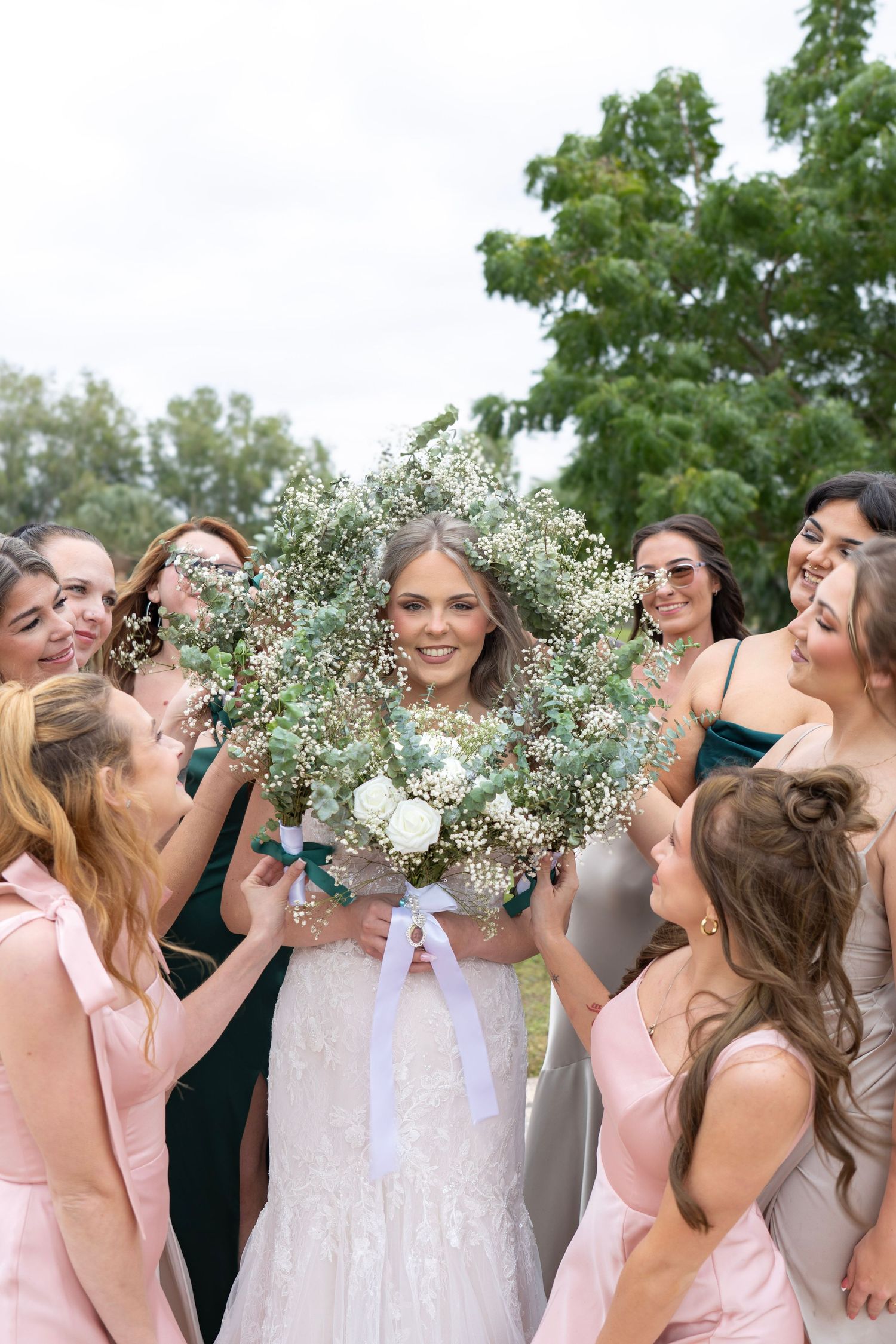 Smiling bride surrounded by bridesmaids in pink and green dresses holding floral wreath bouquet.