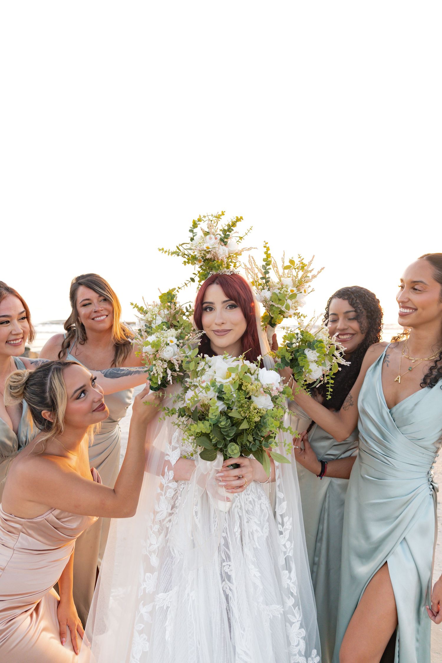 Bride in white lace gown holds floral bouquet surrounded by smiling bridesmaids in sage and blush dresses outdoors.