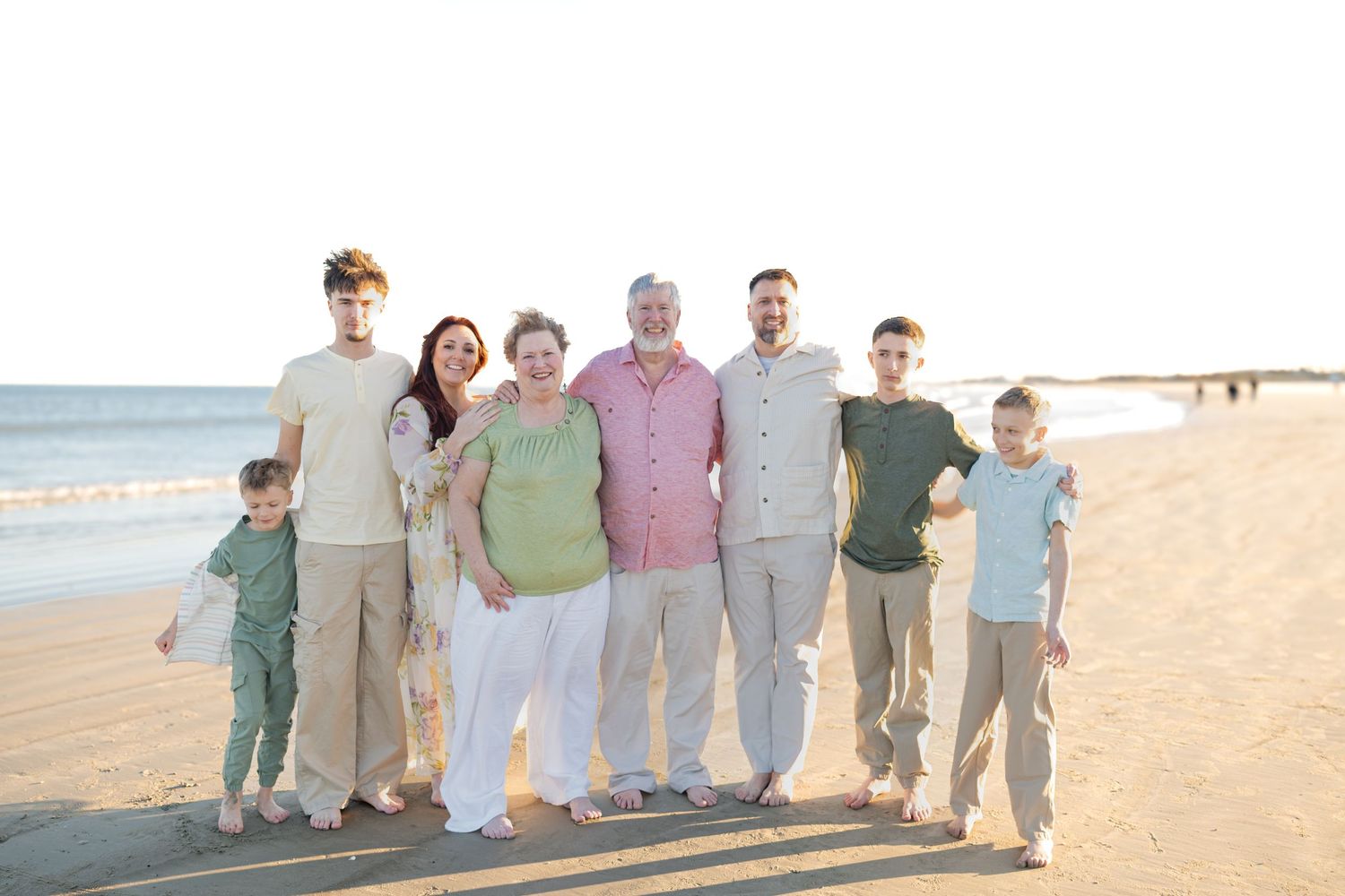 Multi-generational family of eight smiling together on a sandy beach during golden hour.