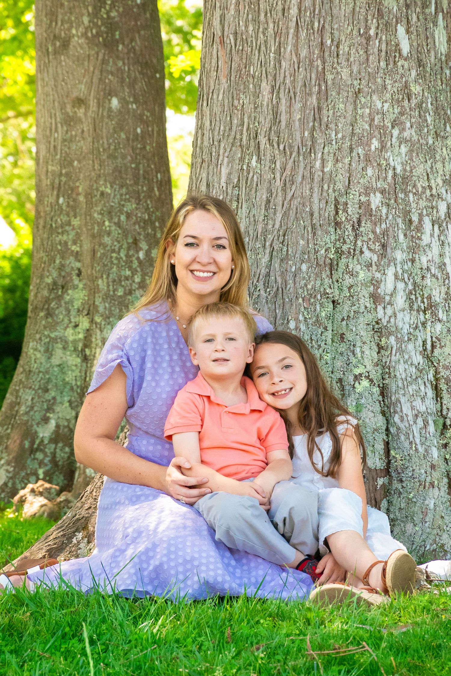 A smiling mother in a purple dress sits with her two young children against a large tree trunk on green grass.