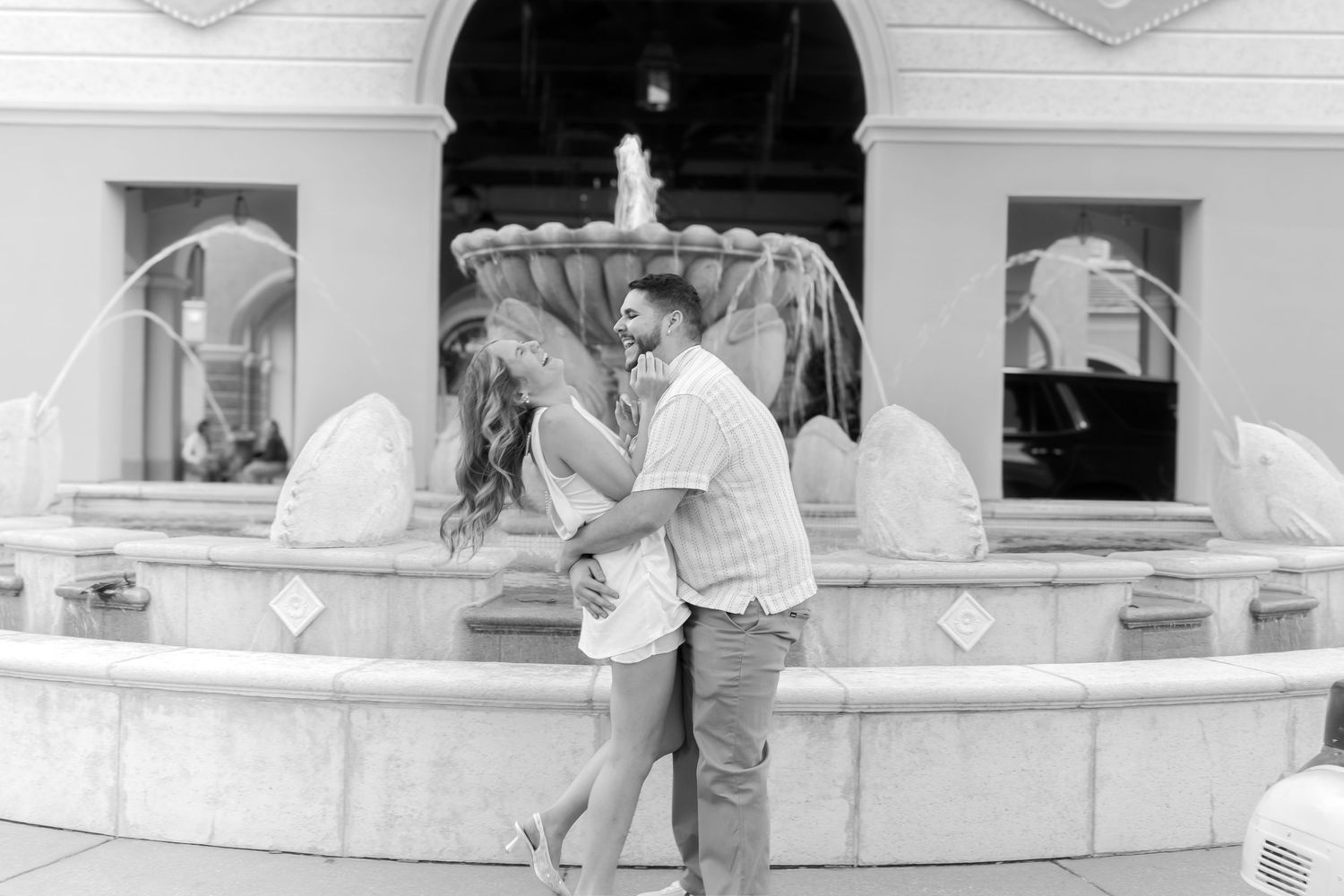 Black and white photo of a couple embracing and kissing in front of an elegant fountain outdoors.