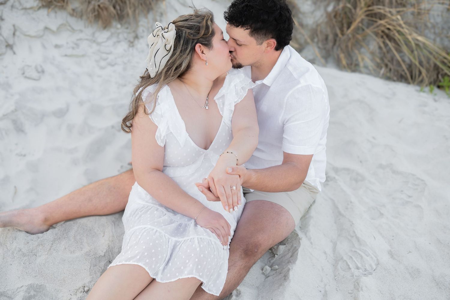Couple sharing a romantic kiss while sitting on white sand dunes, woman in white lace dress, man in white shirt.