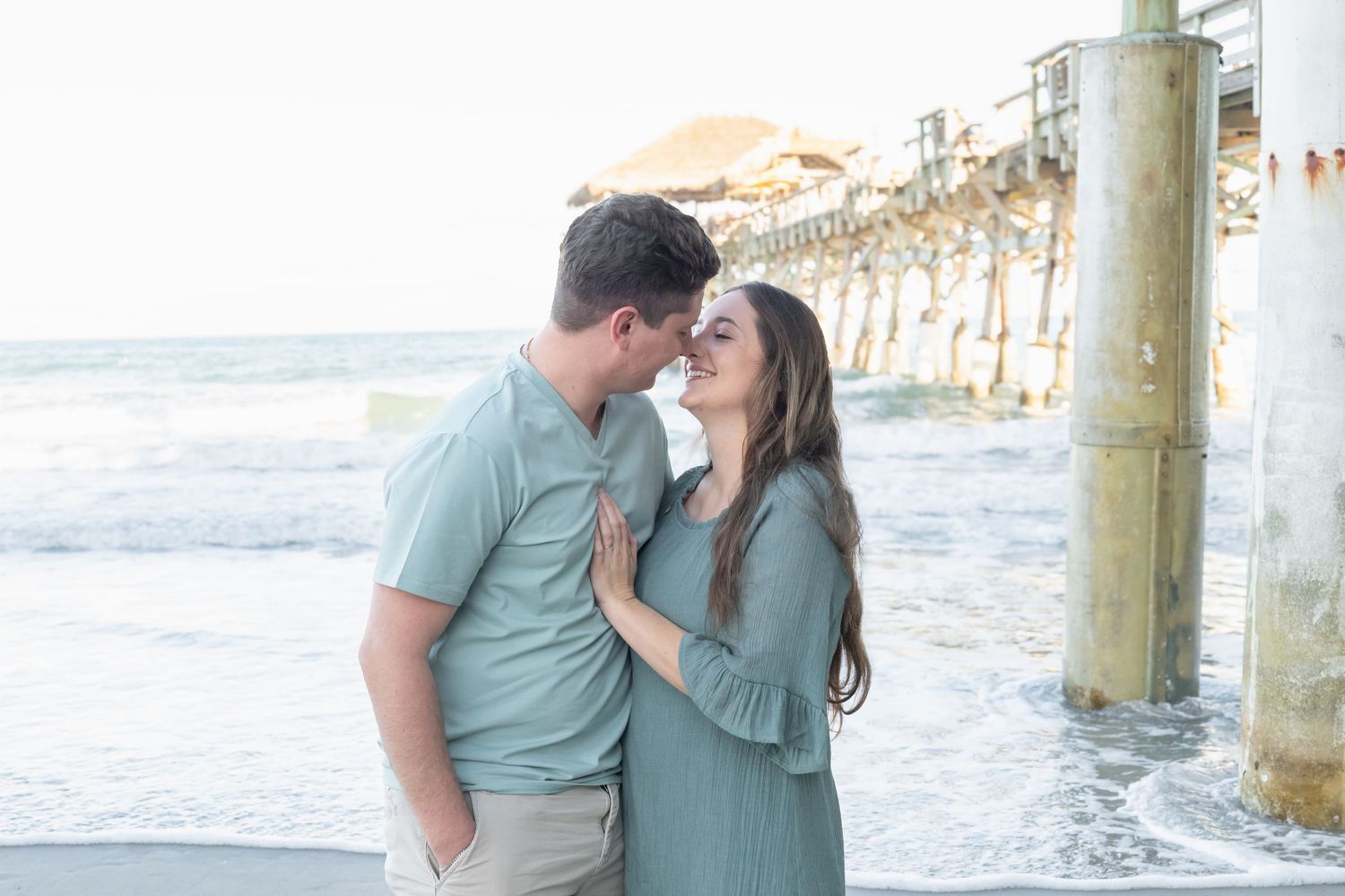 Couple in matching sage green outfits sharing a romantic moment near a beach pier with ocean waves in the background.