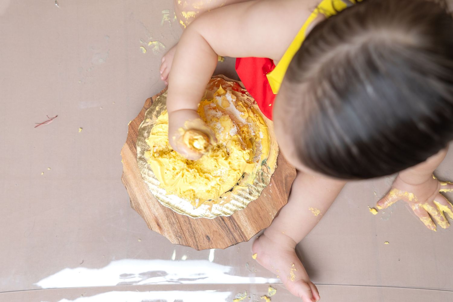 Overhead view of baby smashing yellow birthday cake on wooden board during first birthday cake smash.