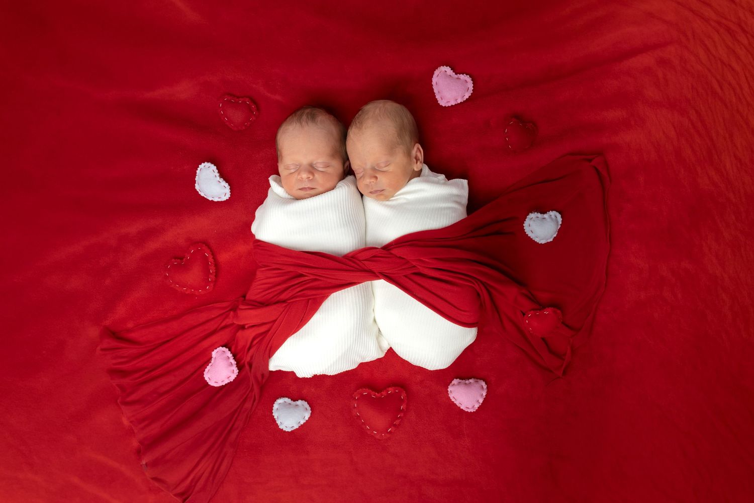 Two swaddled newborn twins sleeping on a red blanket surrounded by heart decorations for Valentine's Day.