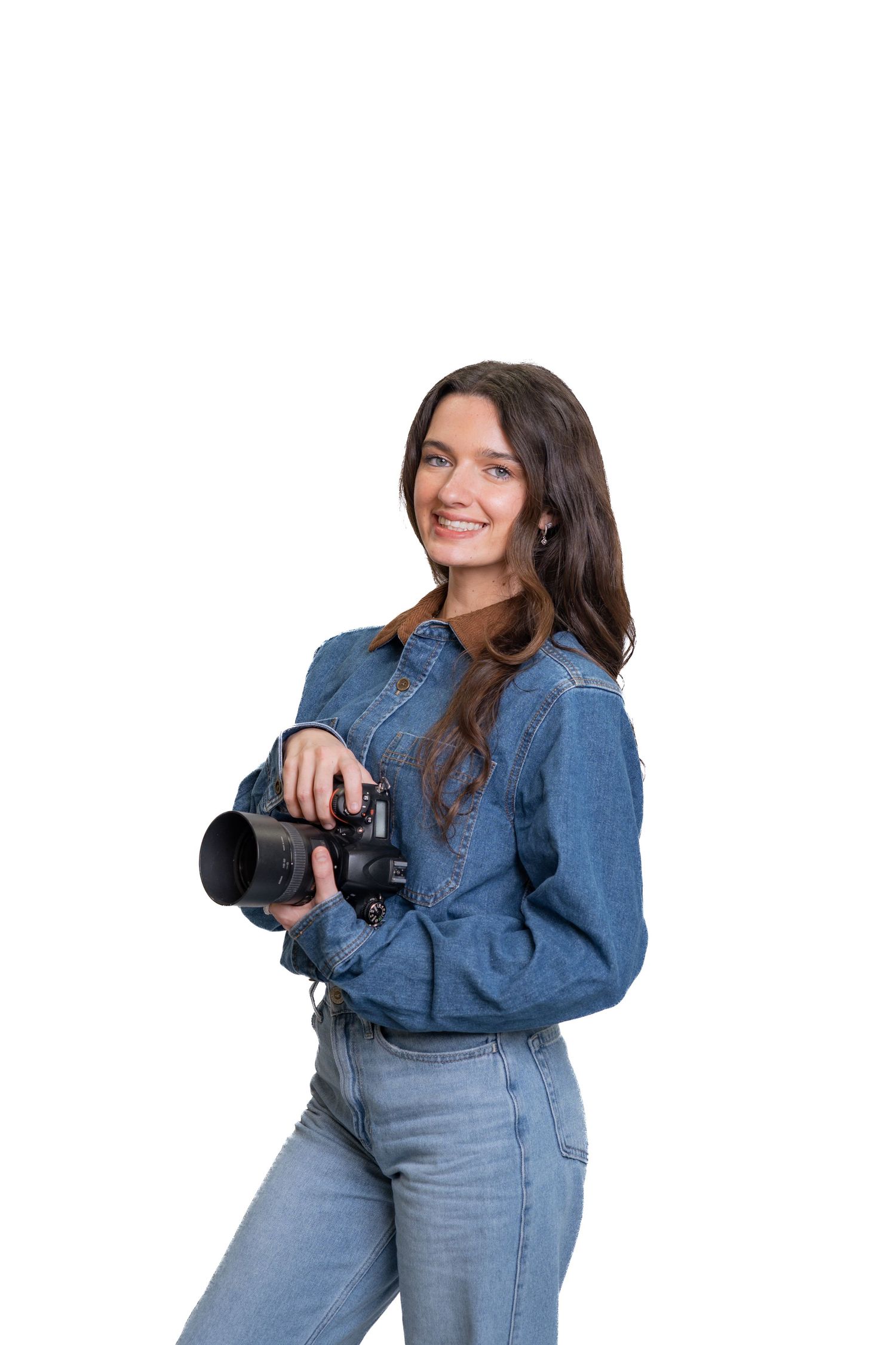 Young female photographer in denim outfit smiling while holding a DSLR camera on a white background.