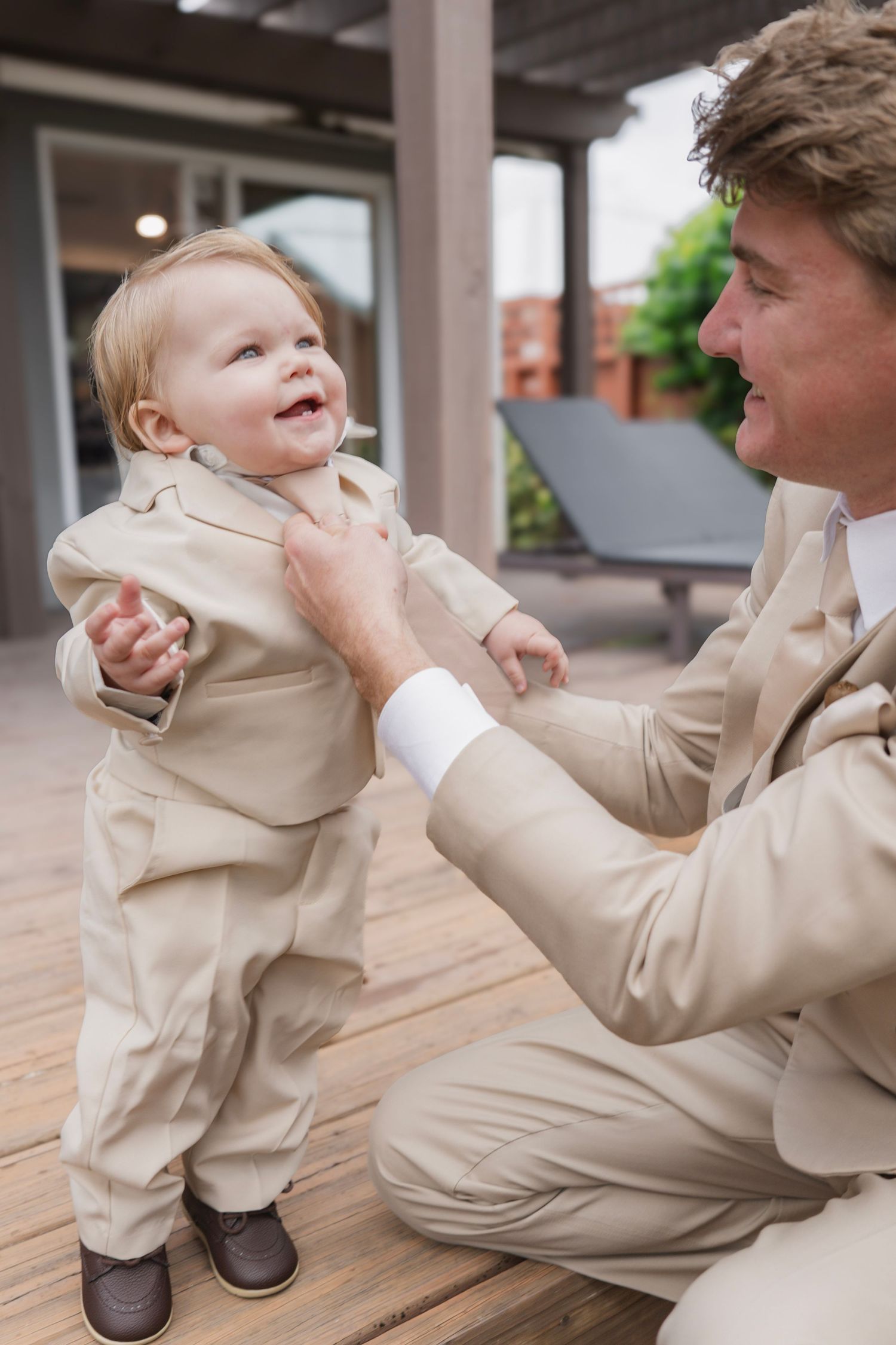 Father in beige suit helping smiling baby boy in matching outfit stand on wooden deck.