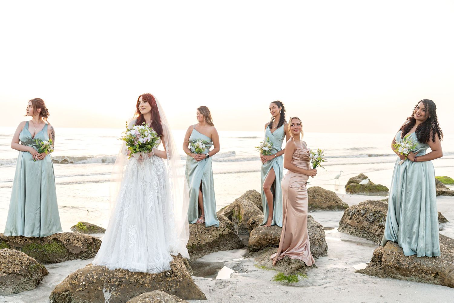 Bride in white gown with bridesmaids in mint and blush dresses posing on rocky beach with ocean backdrop.