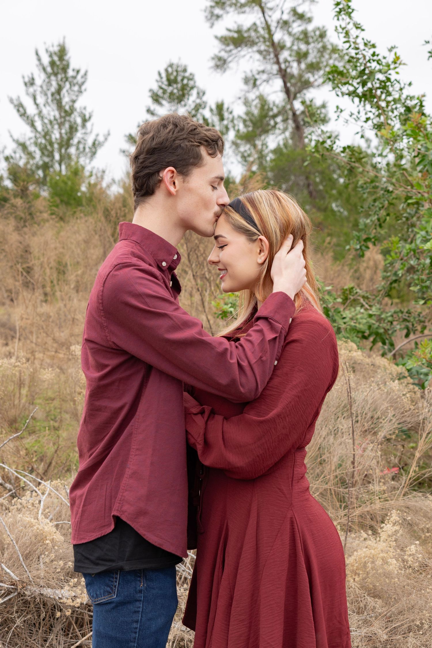 A young couple in matching burgundy outfits embrace tenderly outdoors, surrounded by dry grass and trees.
