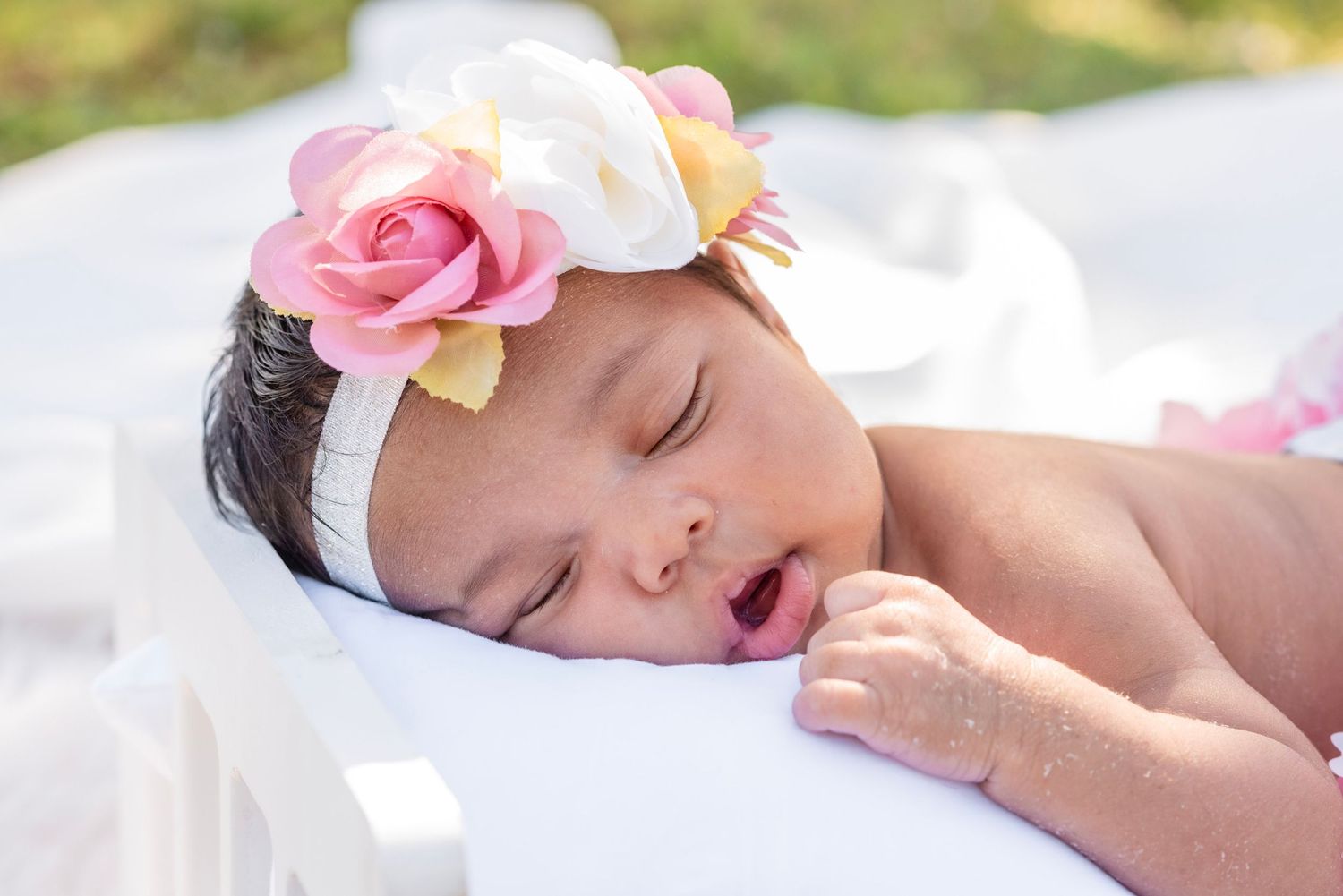 Sleeping newborn baby girl wearing a pink rose floral headband, resting peacefully on a white surface outdoors.
