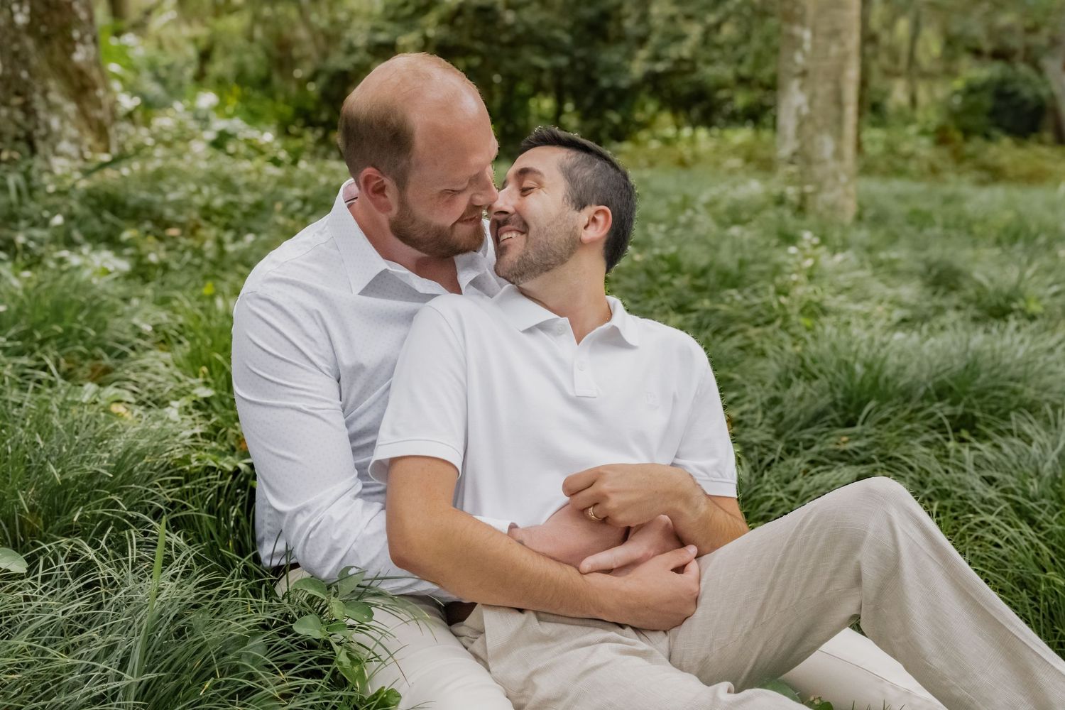 Two men in white shirts sit together in lush green garden, sharing a tender romantic moment outdoors.