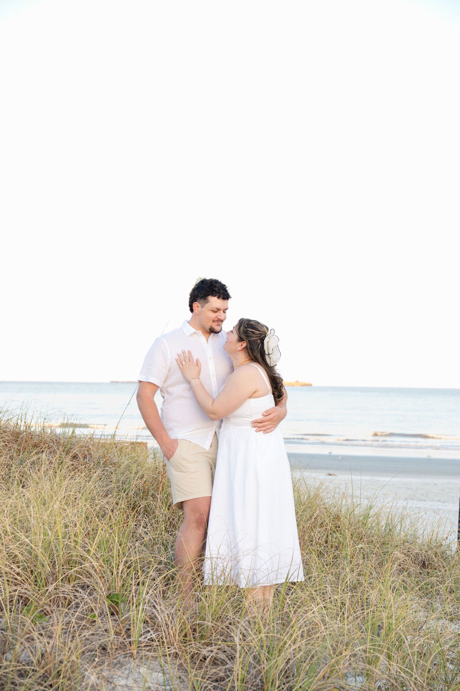 Couple embracing on beach sand dunes, woman in white dress with floral headpiece, man in white shirt and khaki shorts.
