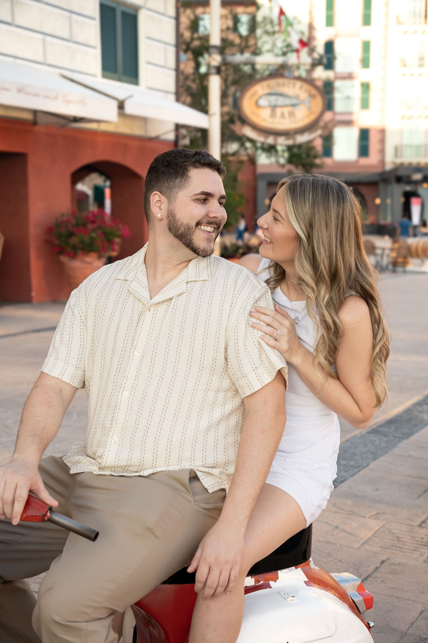 Happy couple laughing together outdoors on a red scooter in a charming town square setting.