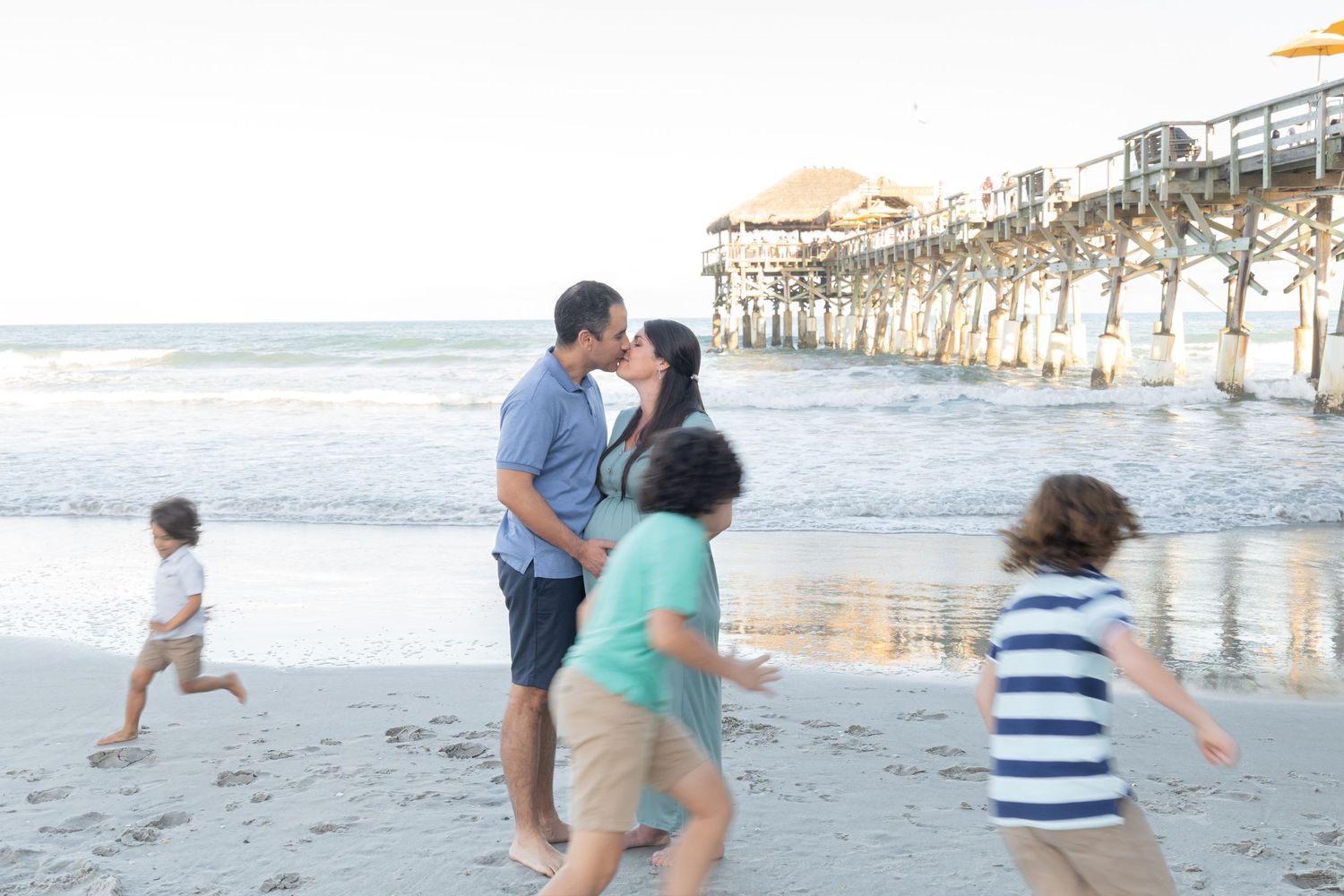 Couple kissing on beach near pier while children play around them at sunset.