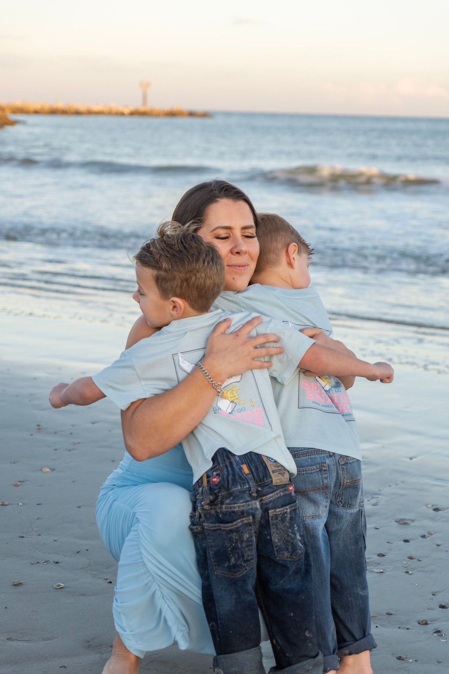 A mother hugging her two young children on a sandy beach at sunset with ocean waves in the background.
