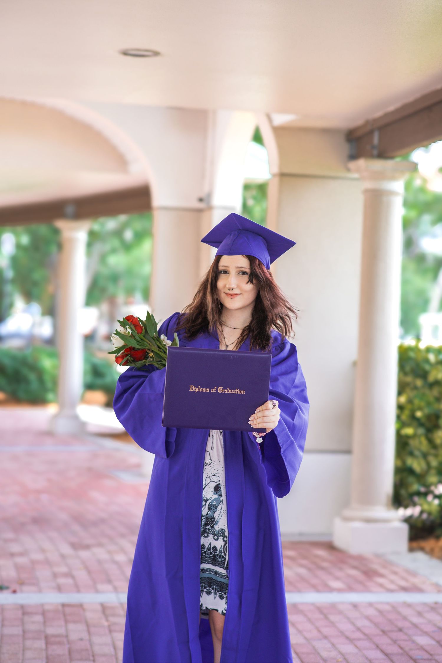 Young woman in purple graduation cap and gown holding diploma and red roses outdoors near white columns.
