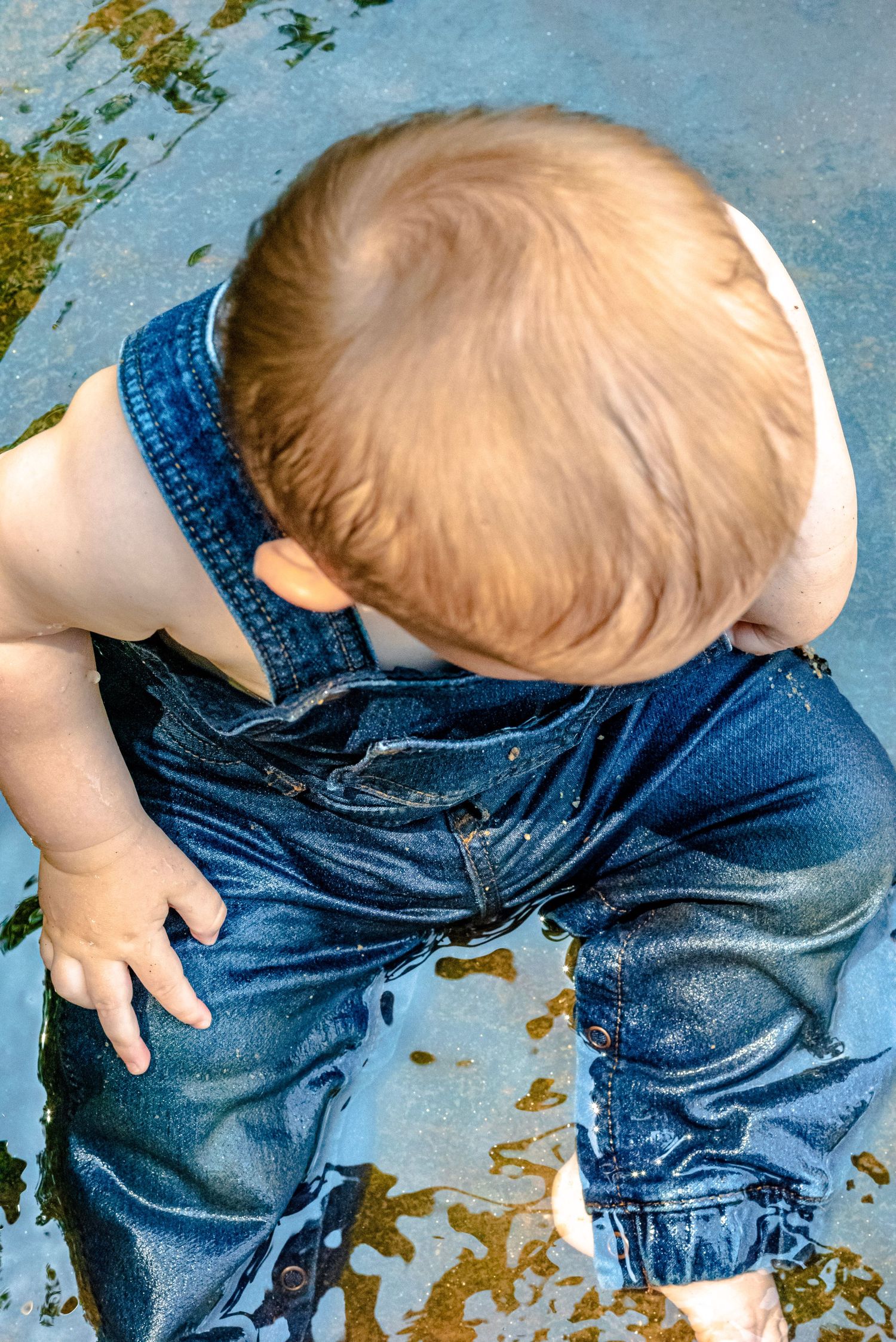 Toddler in wet denim overalls playing in a puddle, viewed from above on a sunny day.