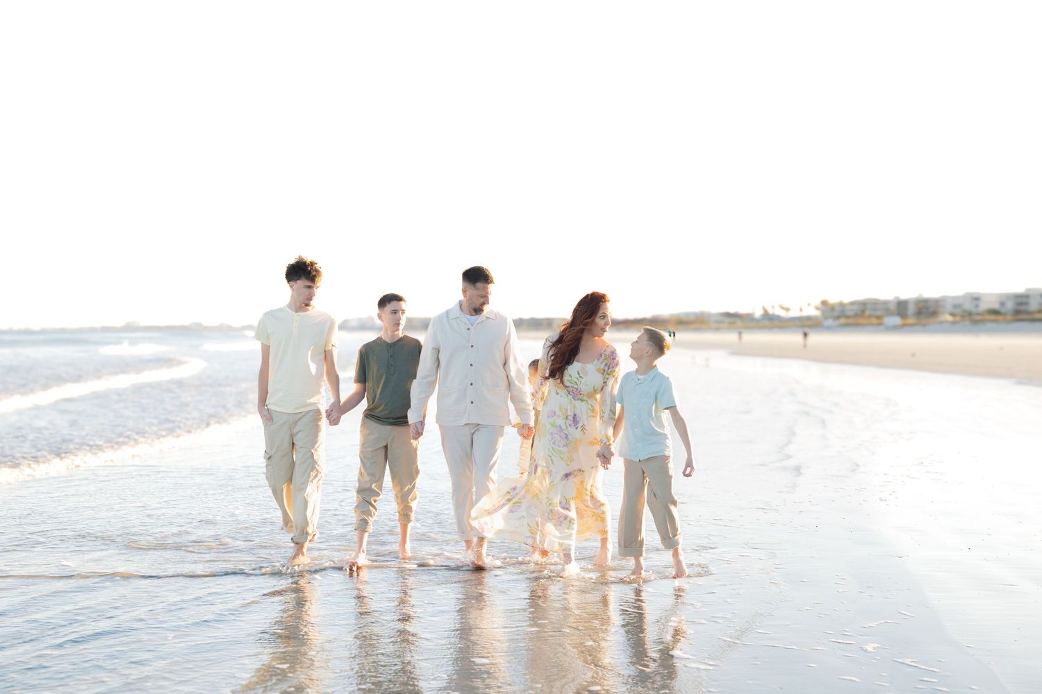 Family of six walking hand-in-hand along a sunlit beach with calm waves and a bright sky.