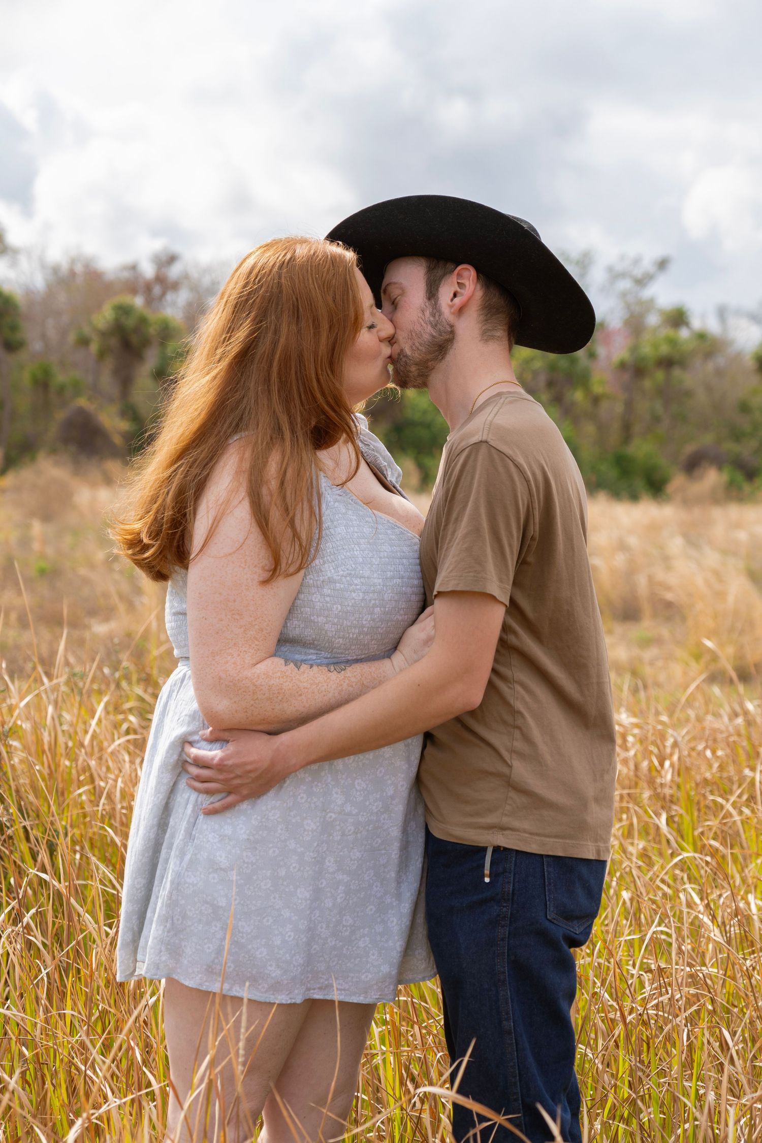 A couple sharing a romantic kiss in a golden field, woman in blue dress with red hair, man in black cowboy hat.