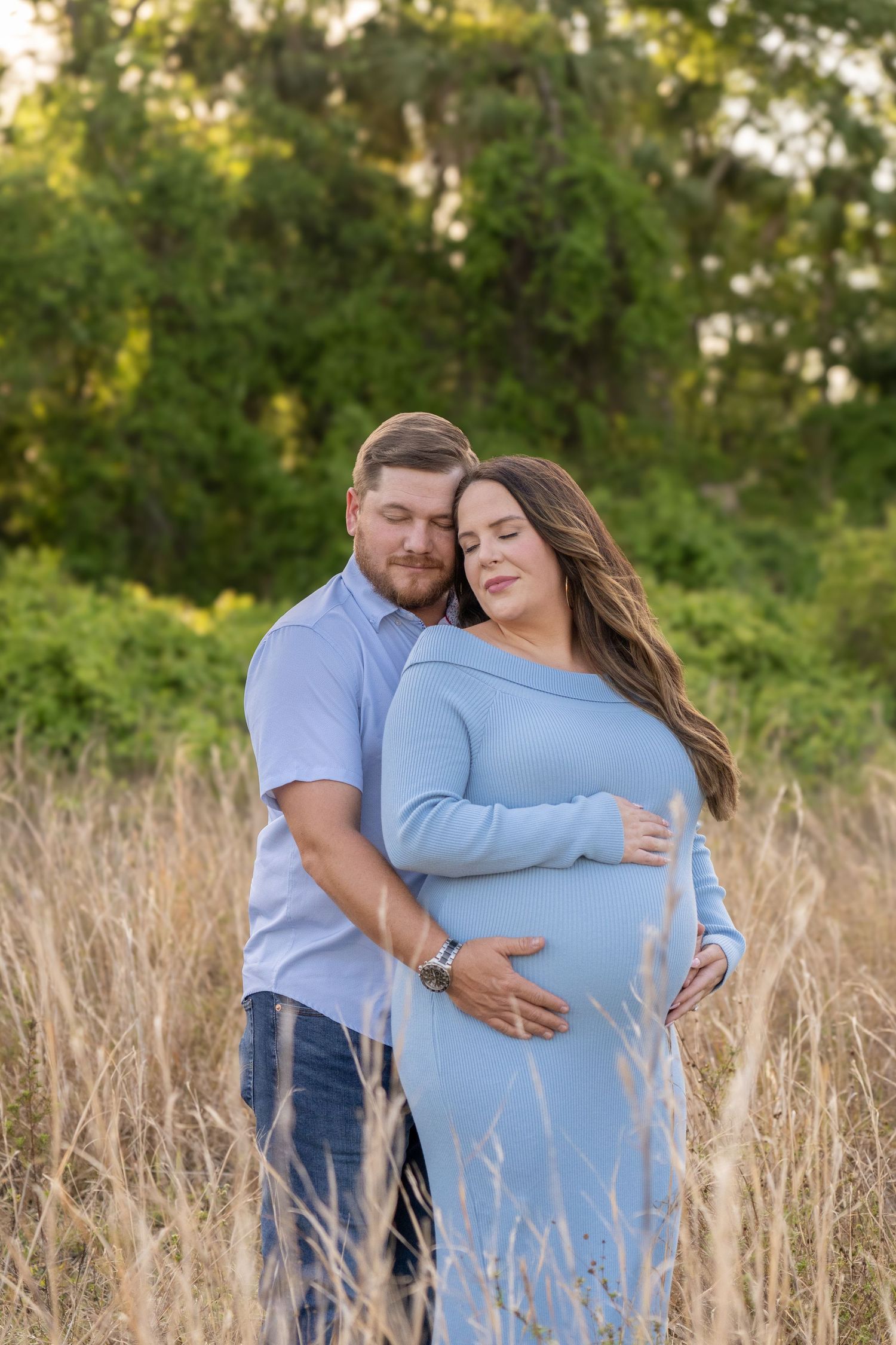 Couple poses in golden field, woman in blue maternity dress cradling baby bump, man embracing her from behind.