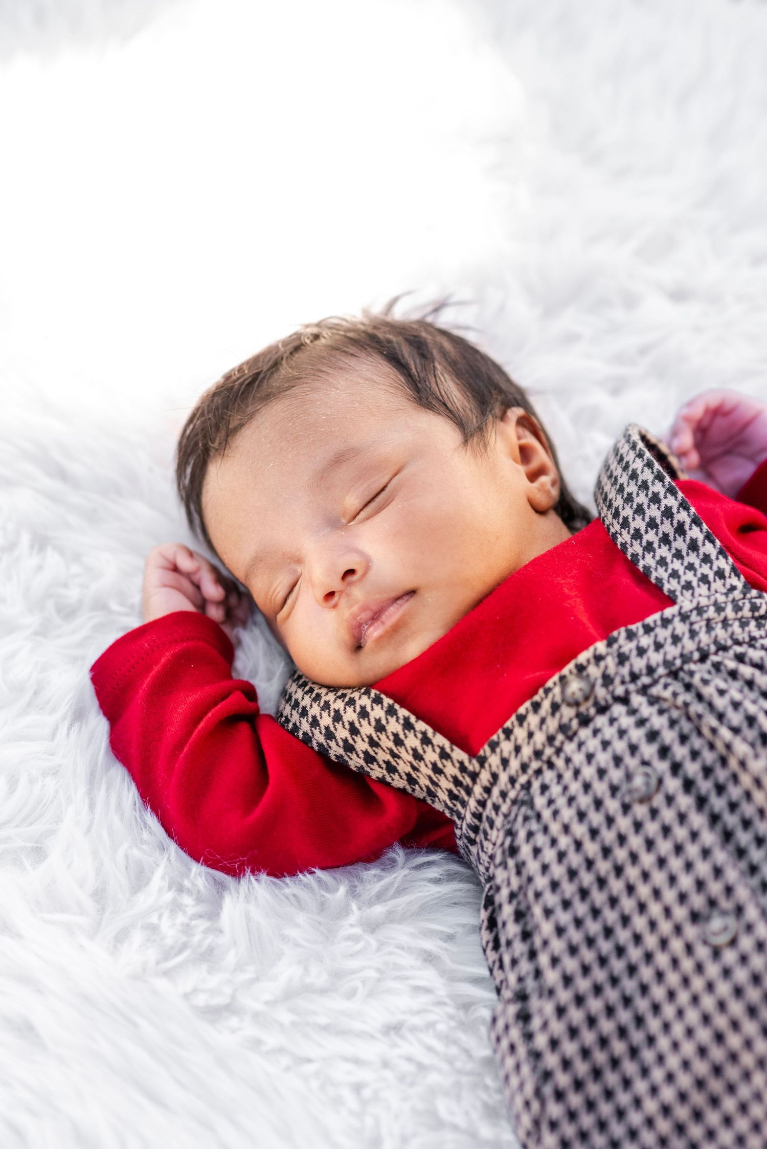 Newborn baby sleeping peacefully on white fluffy blanket wearing red and gray houndstooth outfit.