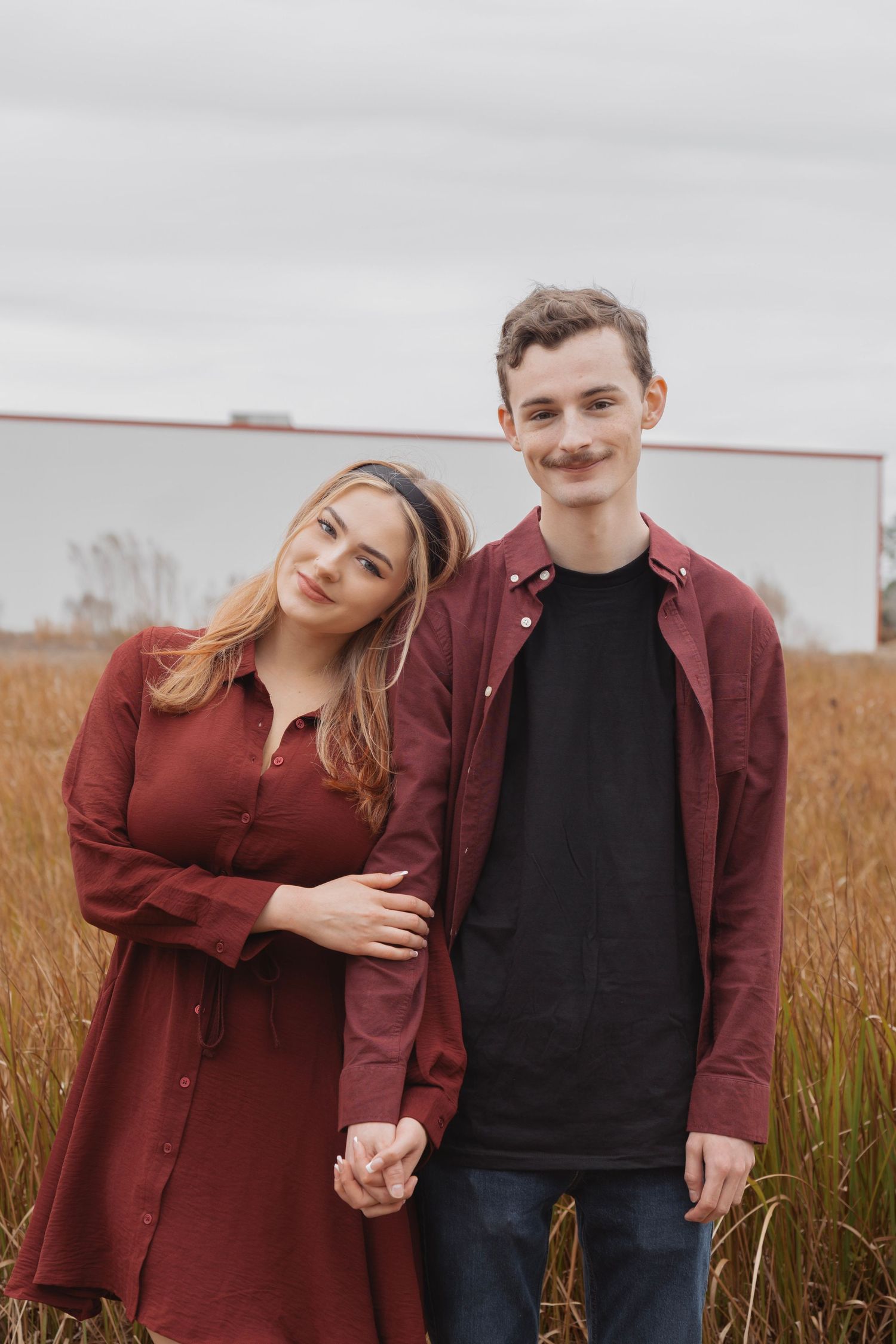 Young couple in matching burgundy outfits holding hands in a golden field on an overcast day.