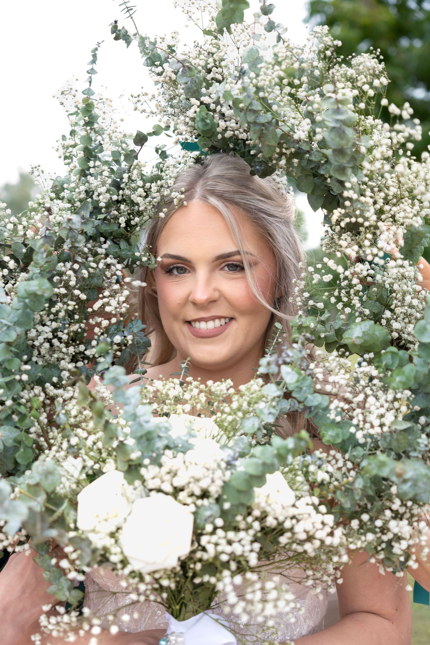 Smiling bride framed by white baby's breath and eucalyptus bouquet, holding white roses at outdoor wedding.