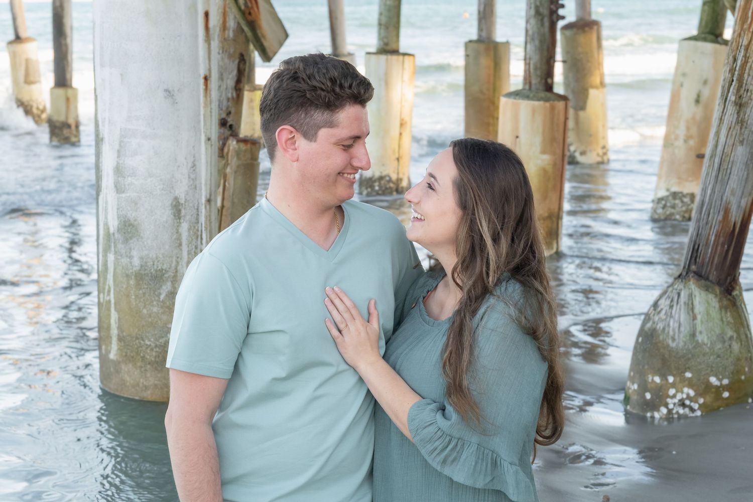 Couple smiling at each other under a beach pier, wearing matching sage green outfits with ocean waves in the background.