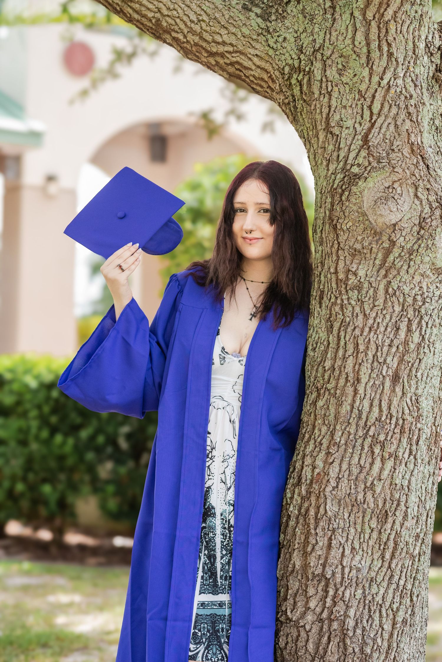 Young woman in blue graduation gown holding cap while leaning against tree on campus.