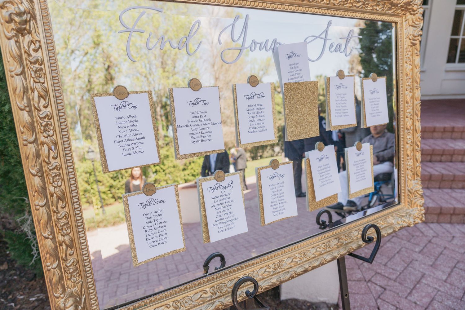 Elegant gold ornate framed wedding seating chart display with white cards on a mirrored background outdoors.