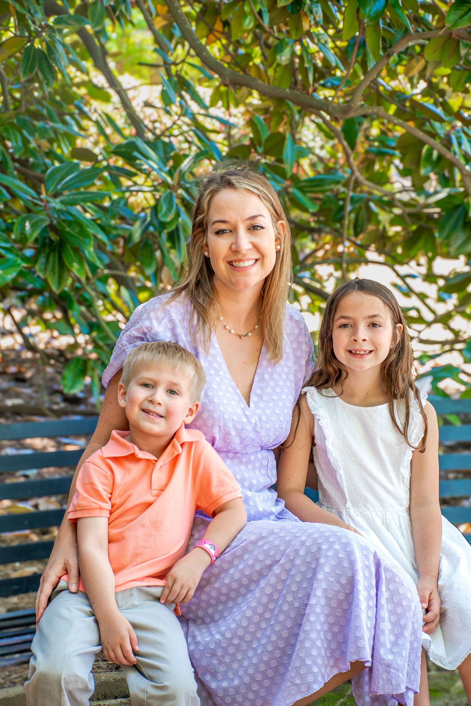 A smiling woman in a purple dress sits on a bench with two young children outdoors near lush green trees.