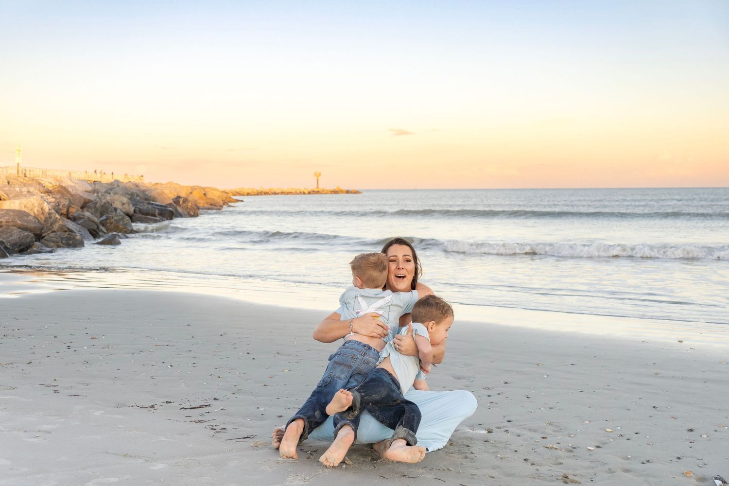 A joyful mother sits on a sandy beach at sunset, laughing while hugging her two young children.