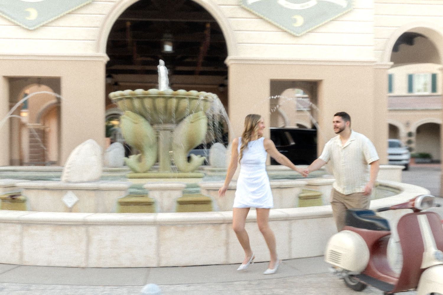 Couple holding hands by ornate fountain with vintage scooter in European-style courtyard.