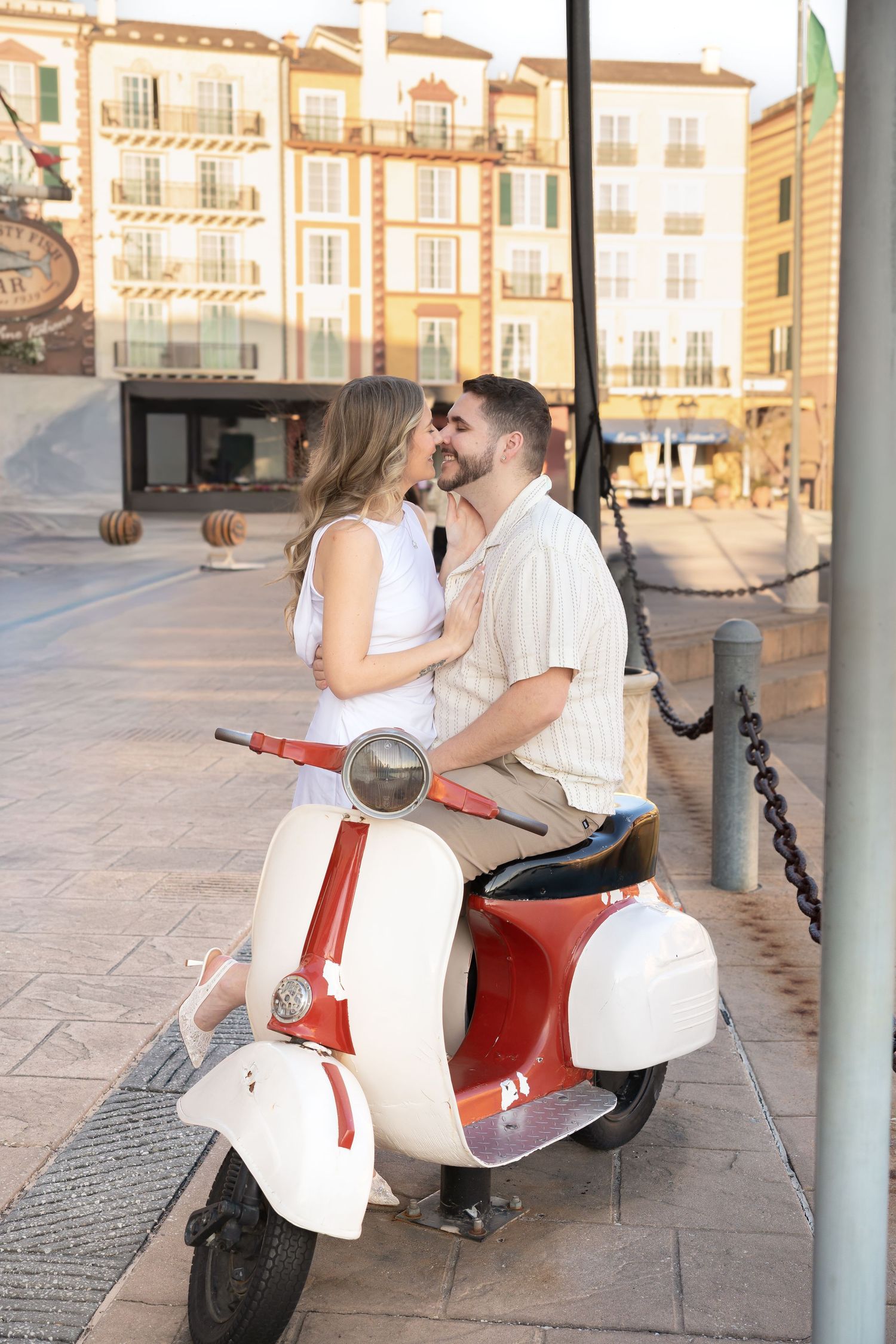 Romantic couple nearly kissing while sitting on a vintage red and white scooter on a sunny plaza.