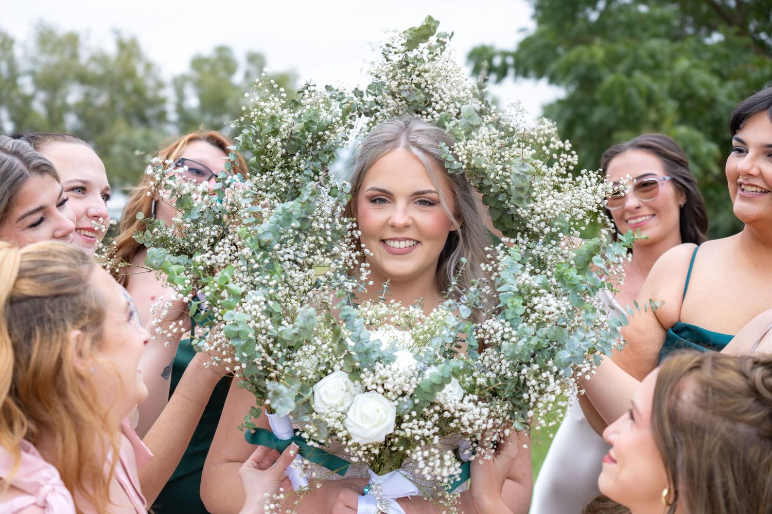 Smiling bride holds a large baby's breath bouquet surrounded by bridesmaids in green dresses outdoors.