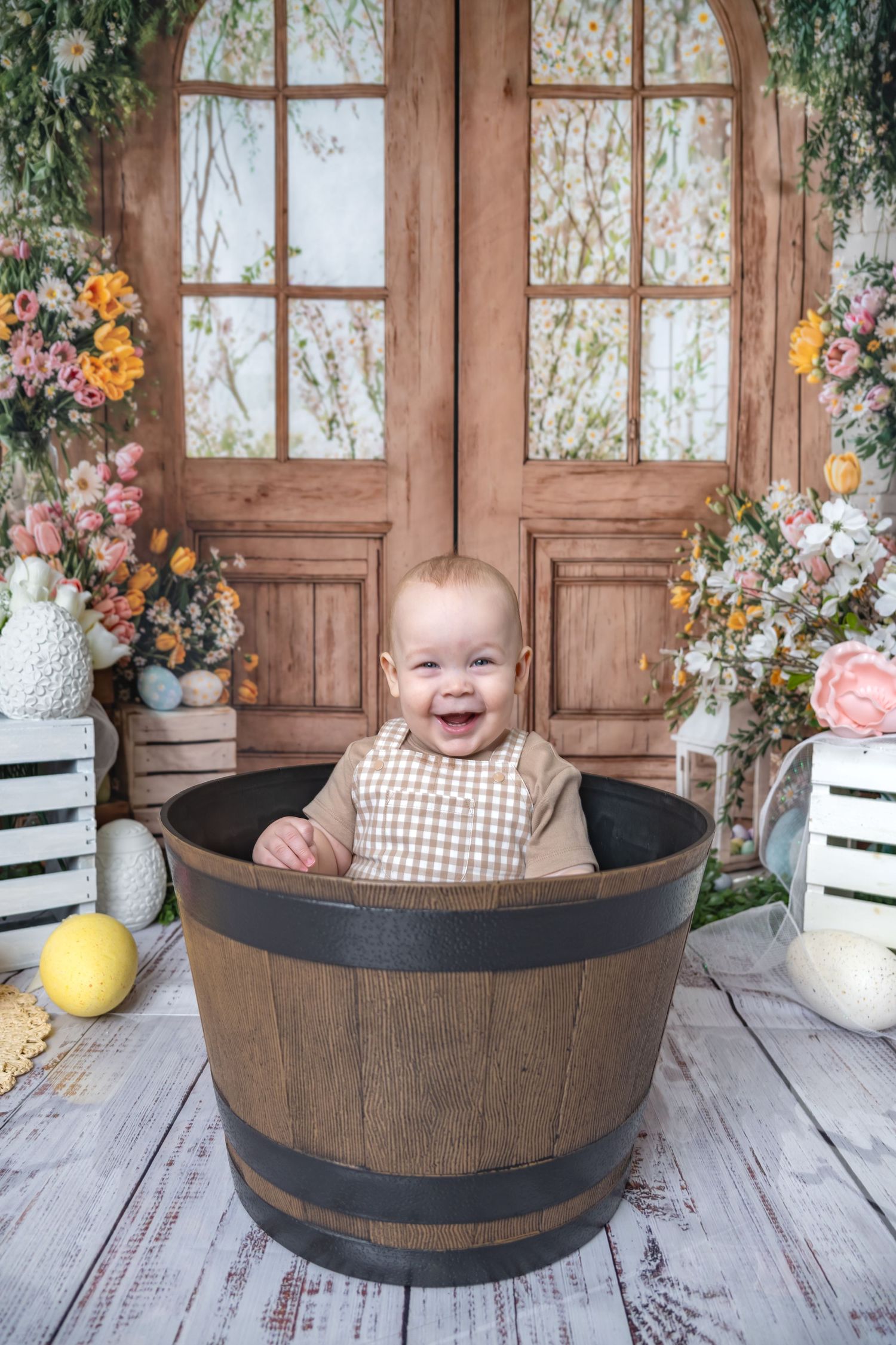 Happy laughing baby sitting in a rustic wooden barrel with a floral spring photography backdrop.