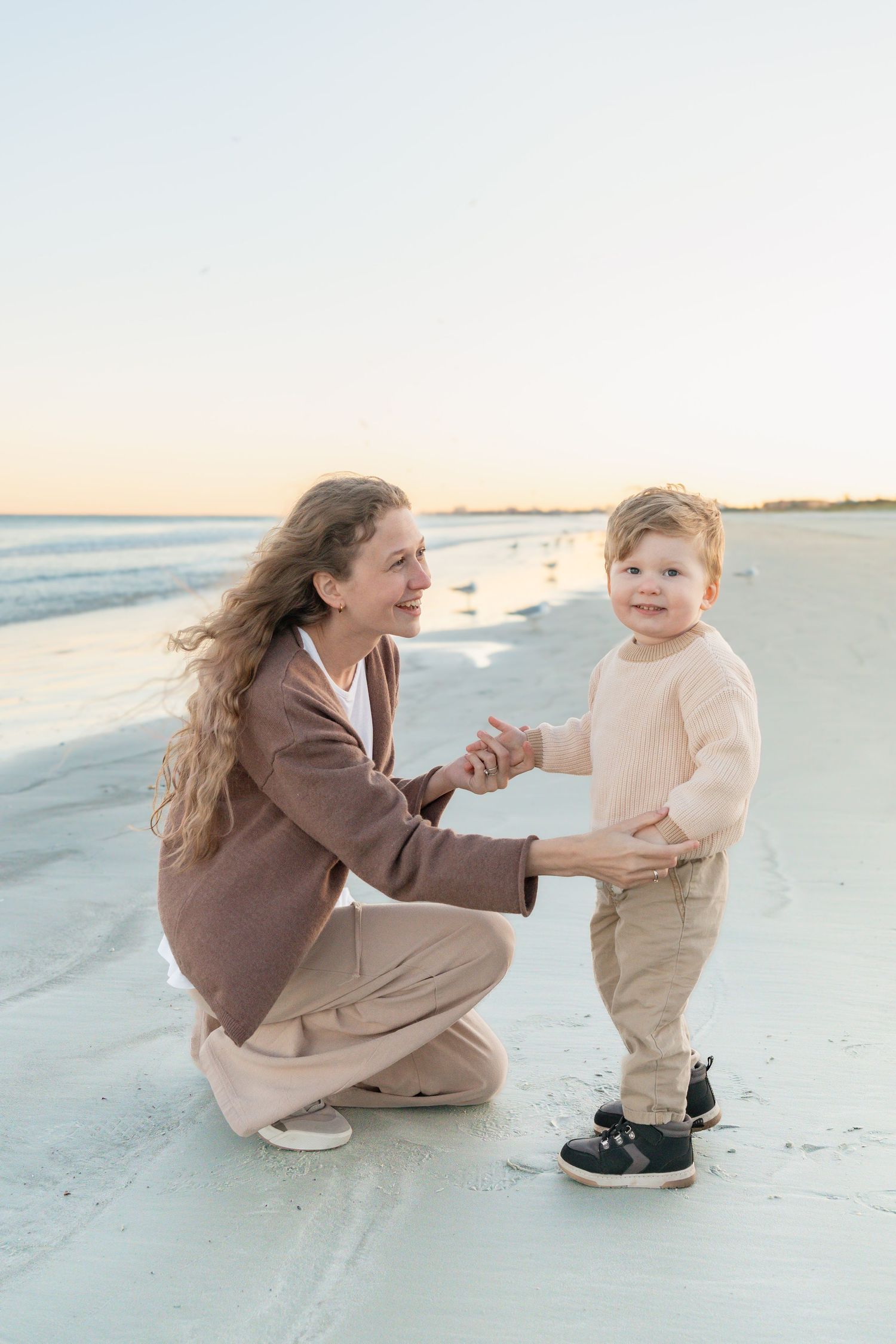 Mother crouching beside her toddler son on a sandy beach at sunset, both smiling and holding hands.