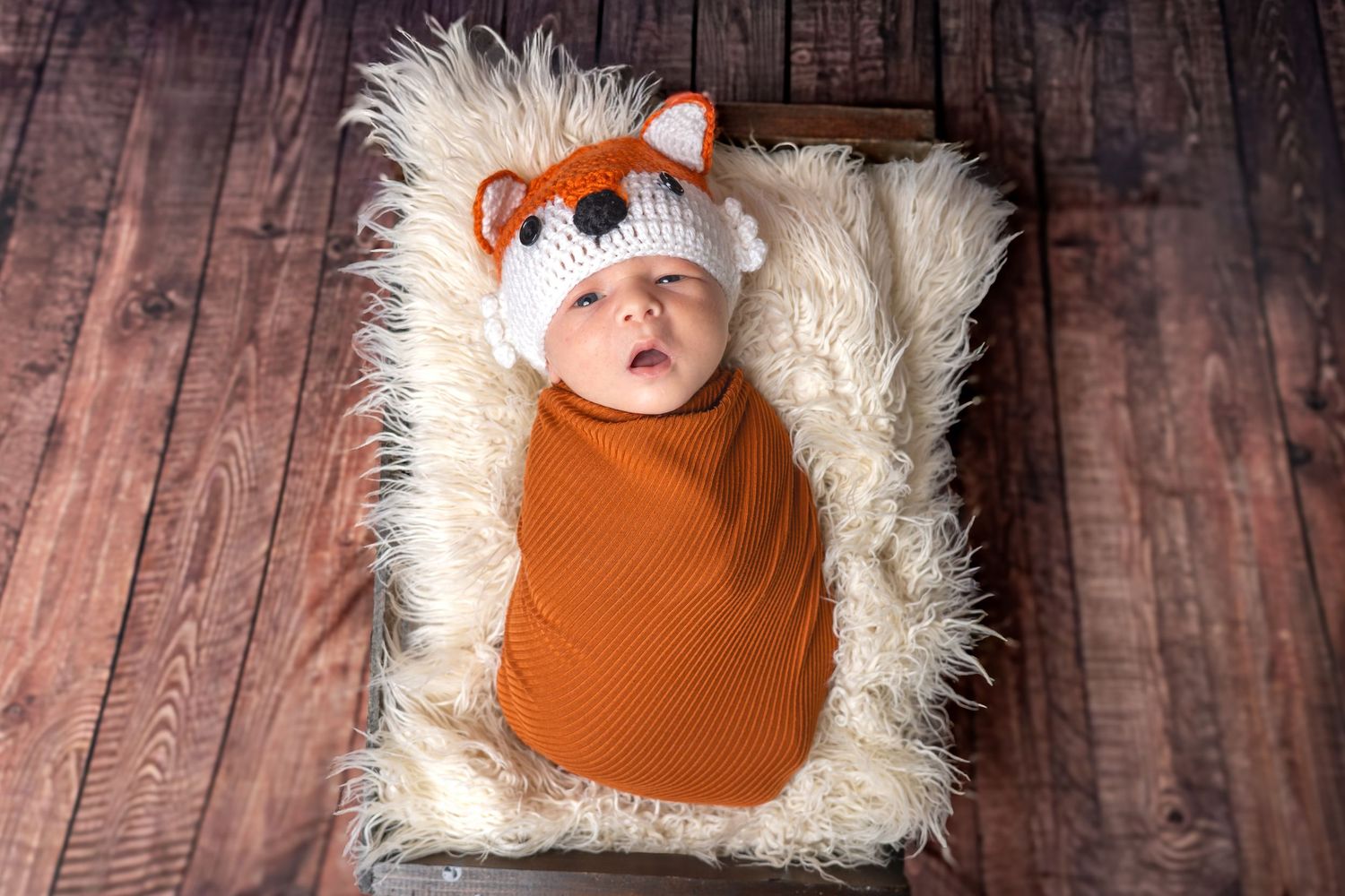 Newborn baby wrapped in orange swaddle wearing a fox knit hat, lying on fluffy white fur in a wooden box.