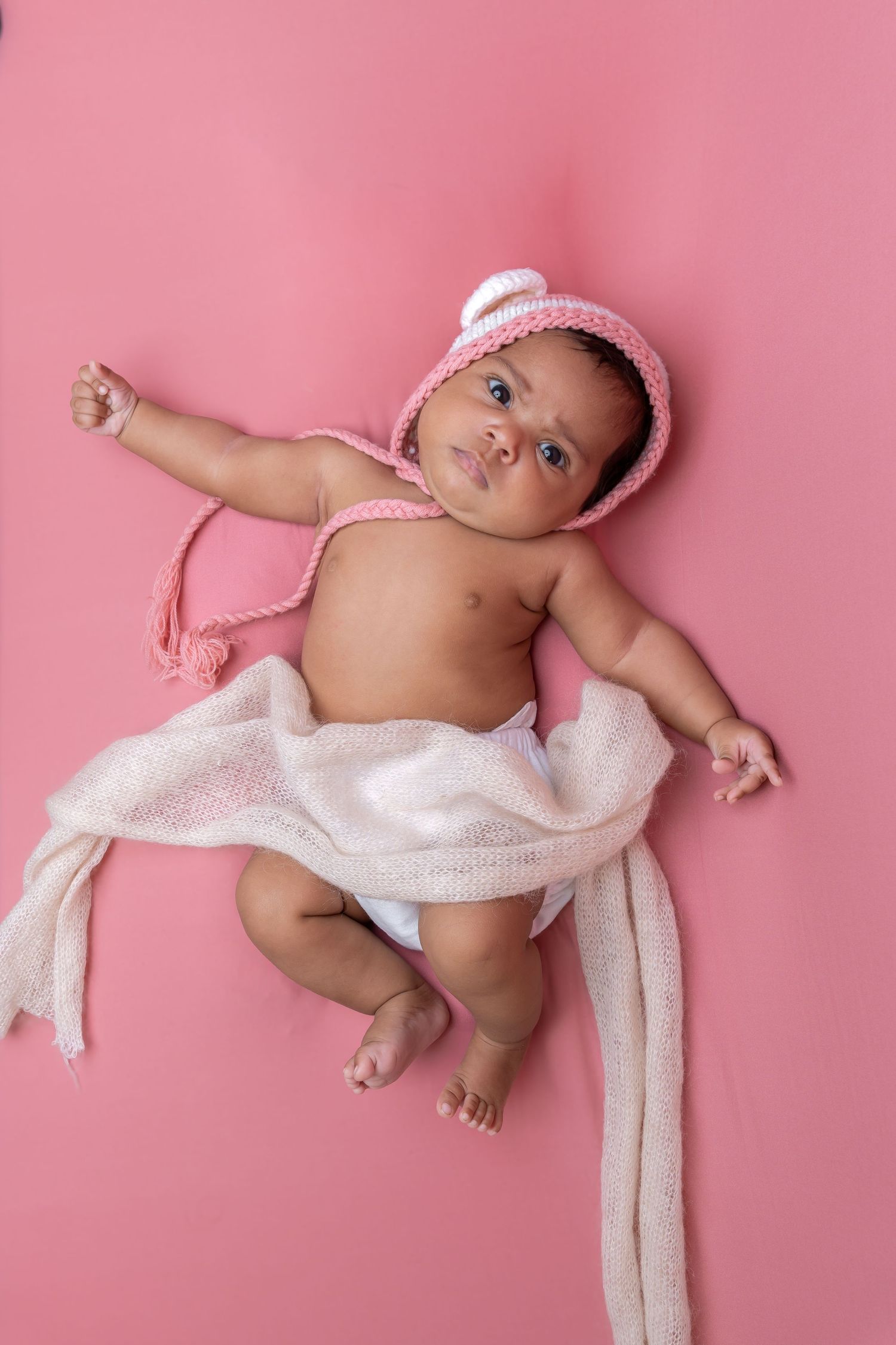 Adorable newborn baby wearing pink crochet hat lying on pink backdrop with white muslin wrap.